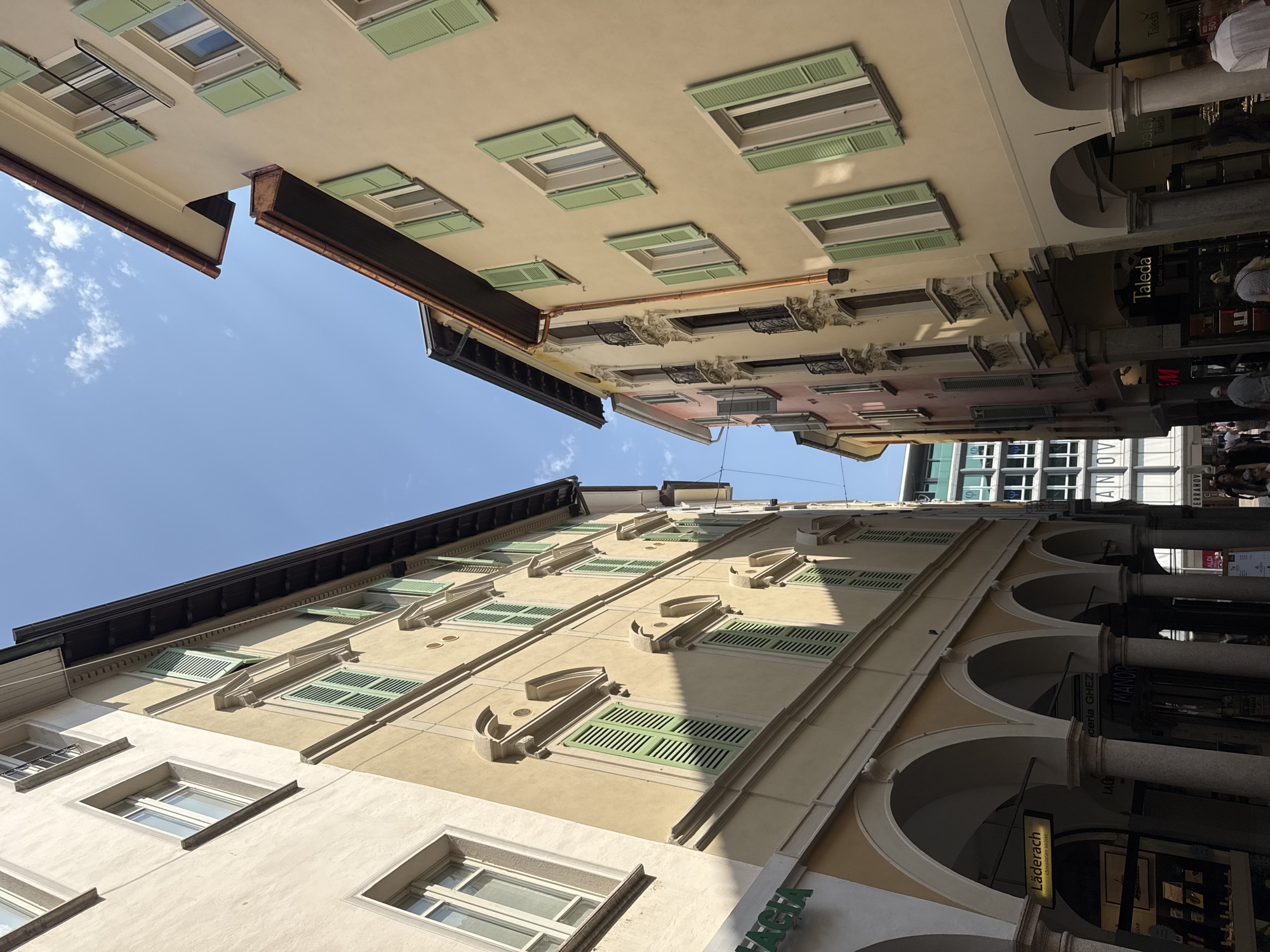 The image shows a narrow street in Lugano, Switzerland, flanked by tall, traditional European buildings. The buildings have light-colored facades, predominantly cream and pale yellow, with numerous windows featuring light green shutters. The architecture suggests a historic European city center, with arcades visible at street level, providing covered walkways for pedestrians and housing shops.
On the left side, an arcade with classical columns supports the upper floors. A sign for "Läderach" (chocolates suisses) is visible, indicating a confectionery shop. The building on this side appears to be older and more ornate.
On the right side, the buildings are also multi-storied with similar window styles and green shutters. The street is paved, and several pedestrians are walking, though their faces are not clearly discernible, suggesting a normal urban scene.
In the background, the sky is bright blue with a few white clouds, indicating clear weather. The buildings are tall enough to create a canyon-like effect in the street. There are also glimpses of more modern architecture in the distance, such as a building with a grid-like facade. The overall impression is a charming, bustling European street scene in Lugano.