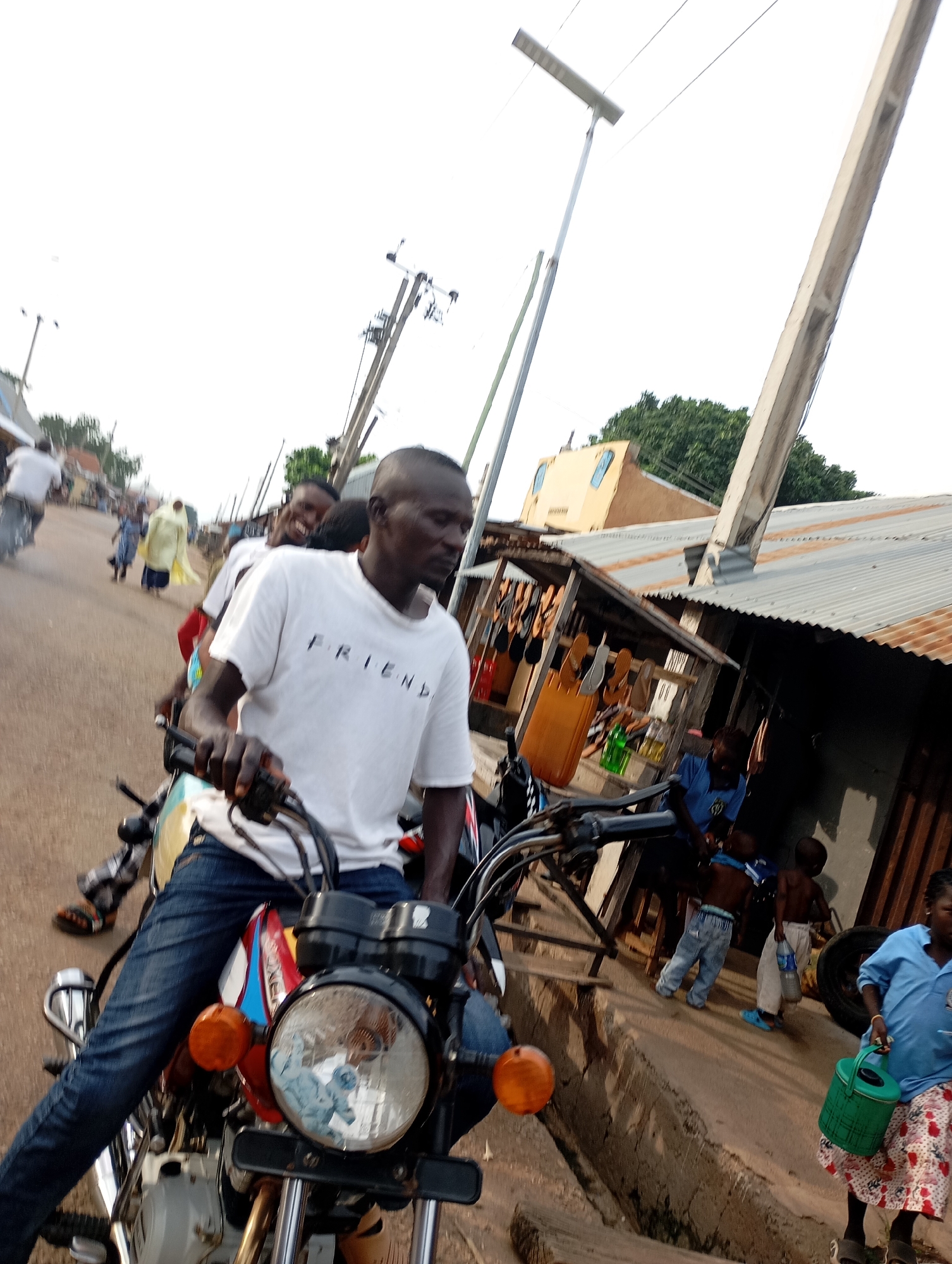 The image depicts a street scene in Shendam, Nigeria, with a focus on a man riding a motorcycle. The man is wearing a white t-shirt with the word "FRIEND" printed on it and blue jeans. He is the primary subject, with his hands on the handlebars and his gaze directed forward. He appears to be in motion, as suggested by the blur of the background and the posture of the rider.
Behind him, another person, likely a passenger, is visible on the motorcycle, with their face partially obscured and a smile evident. Further down the street, several other individuals are present. A person in a bright yellow garment walks away from the camera, and a group of children are seen near some stalls. One child holds a bottle, and another is carrying a green watering can.
The environment includes a dirt or gravel road, with buildings and structures lining the sides. Some of these structures appear to be shops or stalls, with goods displayed. Utility poles and wires are visible in the background, indicating an urban or semi-urban setting. The overall lighting suggests it is daytime, with the sky being bright and potentially overcast. The image conveys a sense of daily life and activity in the depicted location.