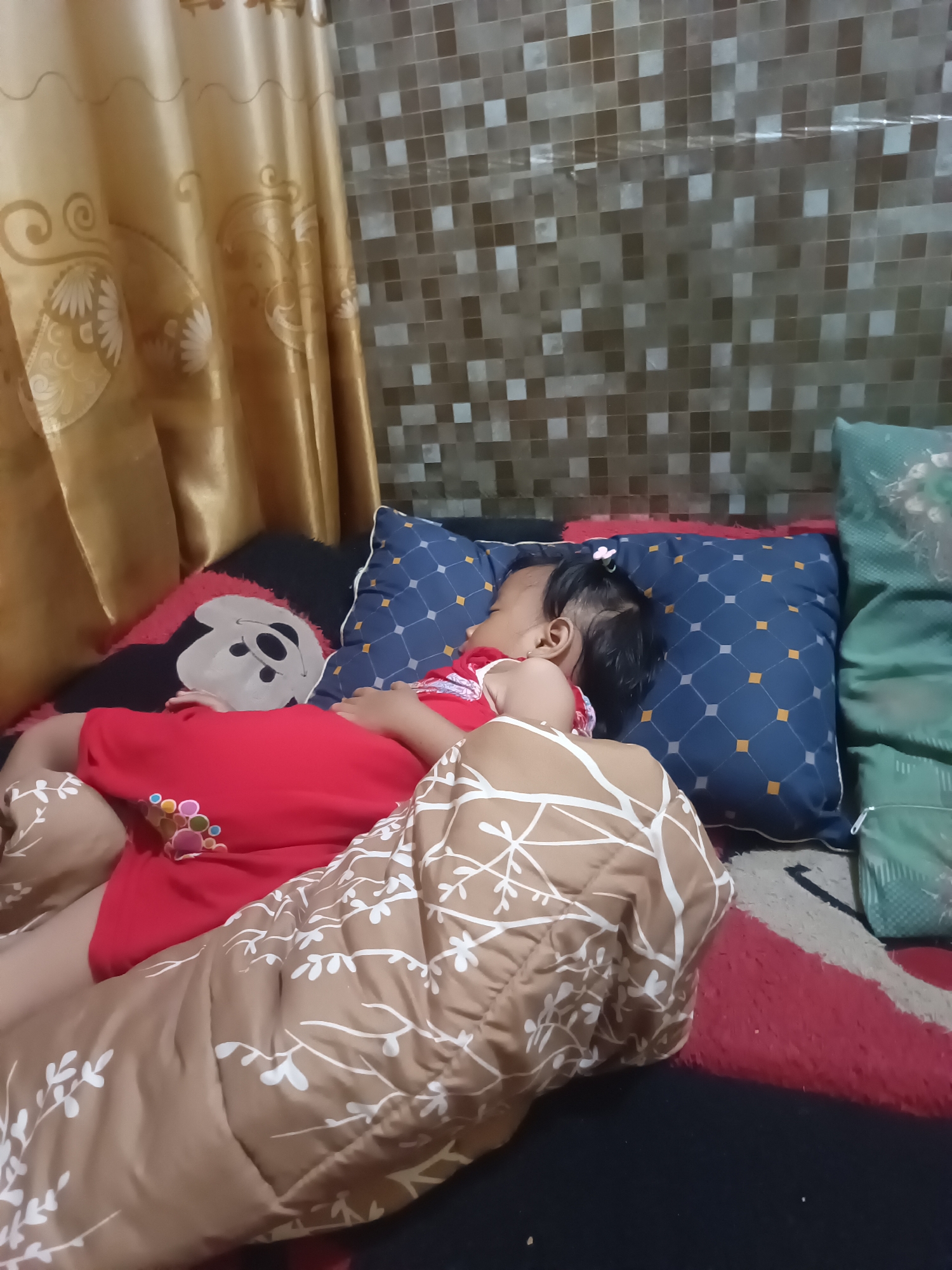 The image depicts a child sleeping on a bed in what appears to be a residential setting. The child, wearing a red shirt with colorful circular embellishments on the front, is lying on their side, covered partially by a brown blanket with a white leafy pattern. Next to the child is a blue pillow with a diamond pattern and a black pillow featuring a Mickey Mouse design. A golden curtain hangs in the background to the left, and a tiled wall is visible behind the pillows. The floor is covered by a rug with red and black sections. The scene is likely indoors, possibly in a bedroom or living area in Pagelaran, Indonesia, given the context.
