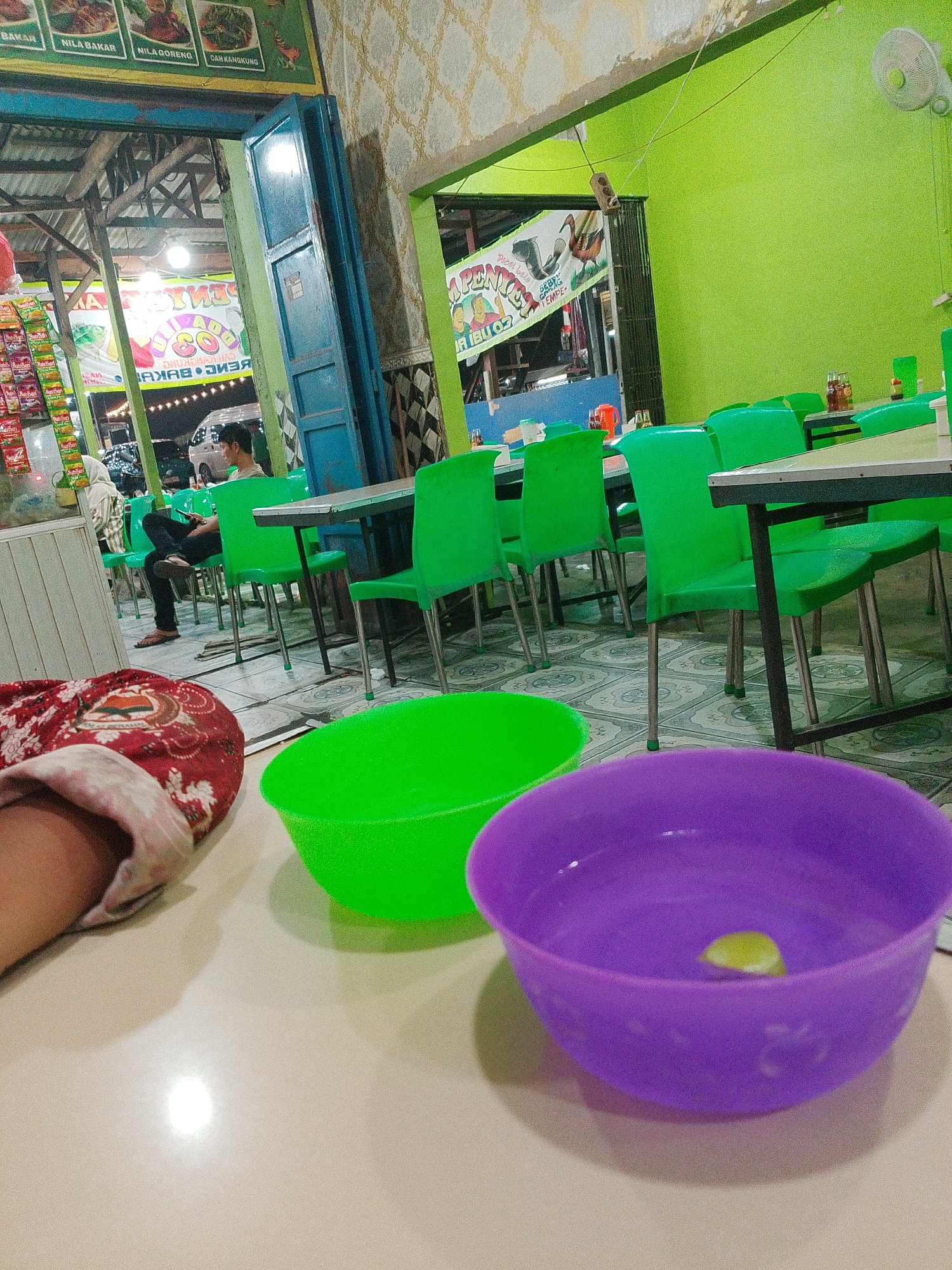The image depicts an indoor dining area, likely a restaurant, with green plastic chairs and tables. A person is seated at one of the tables, looking at their phone. In the foreground, there are two plastic bowls, one green and one purple, on a light-colored table. Behind the tables, a doorway leads to an outdoor area where a person in a headscarf is visible. The walls are decorated with patterned wallpaper and bright green paint. A menu board with images of food is visible on the left side of the image. The overall ambiance suggests a casual dining establishment. The image is taken in Bandar Setia, Indonesia.