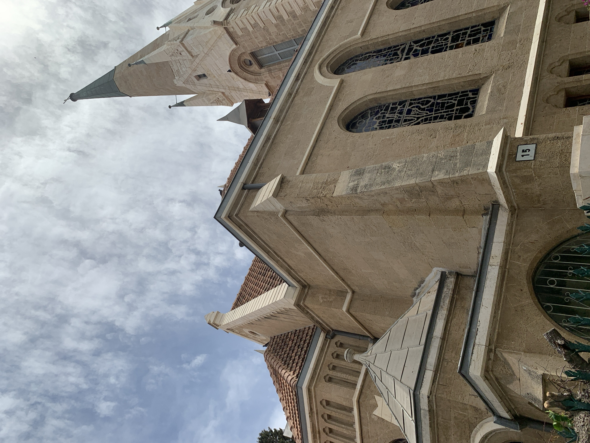 The image shows a low-angle view of a stone building with a pointed tower, possibly a church or similar religious structure. The building has arched windows with decorative metalwork and a roof with red tiles. A sign with the number "15" is visible on the building's wall. The sky is cloudy. Based on the architectural style and the presence of the number 15, this is likely the Immanuel Church in Tel Aviv-Yafo, Israel. No people are visible. The primary focus is on the building's architecture and the surrounding sky.