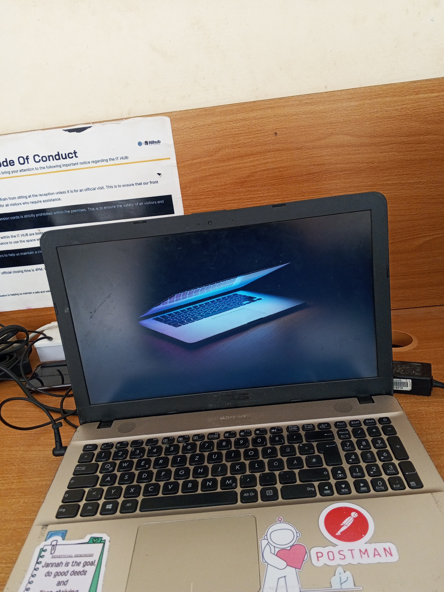 A laptop sits on a desk with a "Code of Conduct" sign nearby, indicating a professional or public space. The laptop screen displays an image of a keyboard against a dark background. The keyboard of the laptop itself is visible, as well as stickers on the base, one with the text "POSTMAN" and another with the text "Jannah is the goal, do good deeds". The environment appears to be an indoor workspace with a wooden desk surface. Based on the Code of Conduct signage that features the name Nihub, and the location metadata included in the prompt, the image was taken at Nihub in Baganakwo, Nigeria.