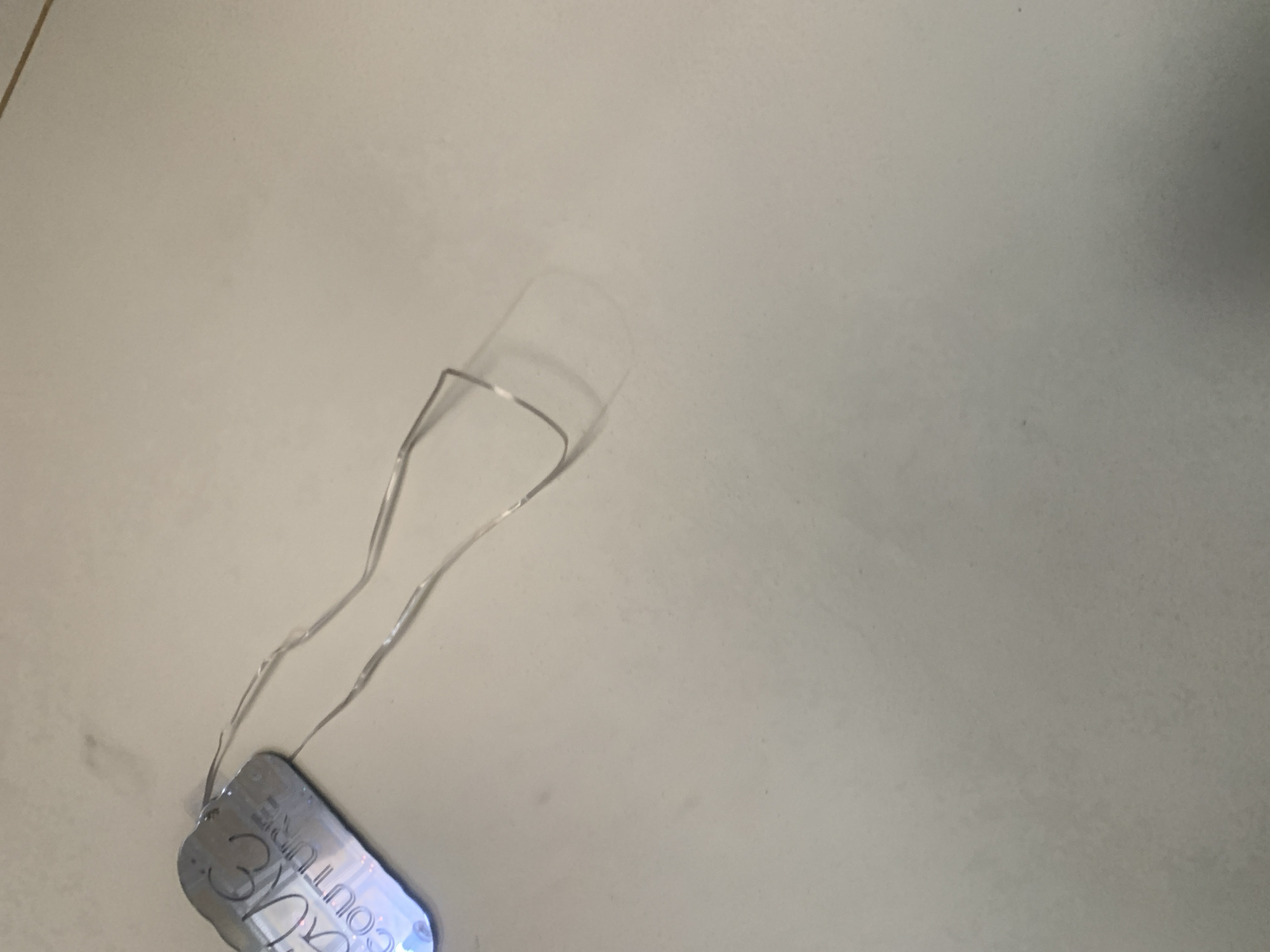 An overhead shot shows a metallic tag with the words "AG COUTURE" etched into it. The tag is attached to a thin, silver string that has been bent into a loop. The background is a neutral, light-colored surface, possibly a countertop or floor. There are no people or other identifiable objects in the scene.