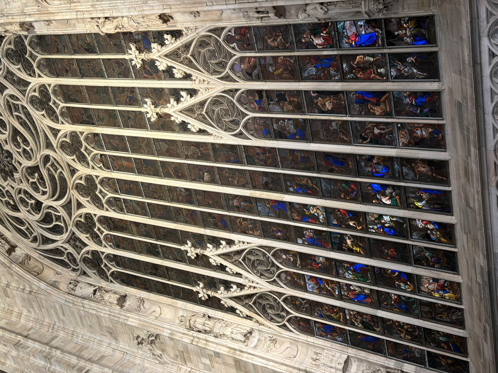 This image presents a dramatic, upward-angled view of a section of a grand Gothic cathedral interior, strongly suggesting the Milan Cathedral given the provided context. The primary subjects are a series of intricate stained glass windows set within elaborate stone tracery, flanked by ornate architectural elements.
The stained glass windows are arranged in multiple horizontal rows, each panel depicting distinct scenes with numerous figures. These figures, adorned in robes, are rendered in various dynamic poses, with prominent colors like deep blues, rich reds, yellows, and earthy browns, contrasting against darker backgrounds. The scenes appear to be biblical or religious narratives, possibly depicting saints, apostles, or events from the life of Christ.
Below and to the left of the stained glass, magnificent Gothic stone tracery forms complex geometric and floral patterns, creating a delicate yet robust framework for the windows. This tracery features pointed arches, quatrefoils, and other intricate designs, characteristic of high Gothic architecture. Further down and to the left, robust stone architecture includes pilasters and decorative elements with relief carvings and what appear to be small statues or figures set in niches, further enhancing the grandeur of the structure.
The image is captured from an elevated, tilted perspective, looking up towards the ceiling or upper wall, emphasizing the immense scale and height of the cathedral. The lighting suggests an interior setting, possibly with natural light filtering through the stained glass or artificial illumination, highlighting the vibrant colors of the glass against the more subdued tones of the stone. No visible text is present in the image. The overall impression is one of awe-inspiring architectural and artistic mastery, characteristic of a major European cathedral like the Duomo di Milano.