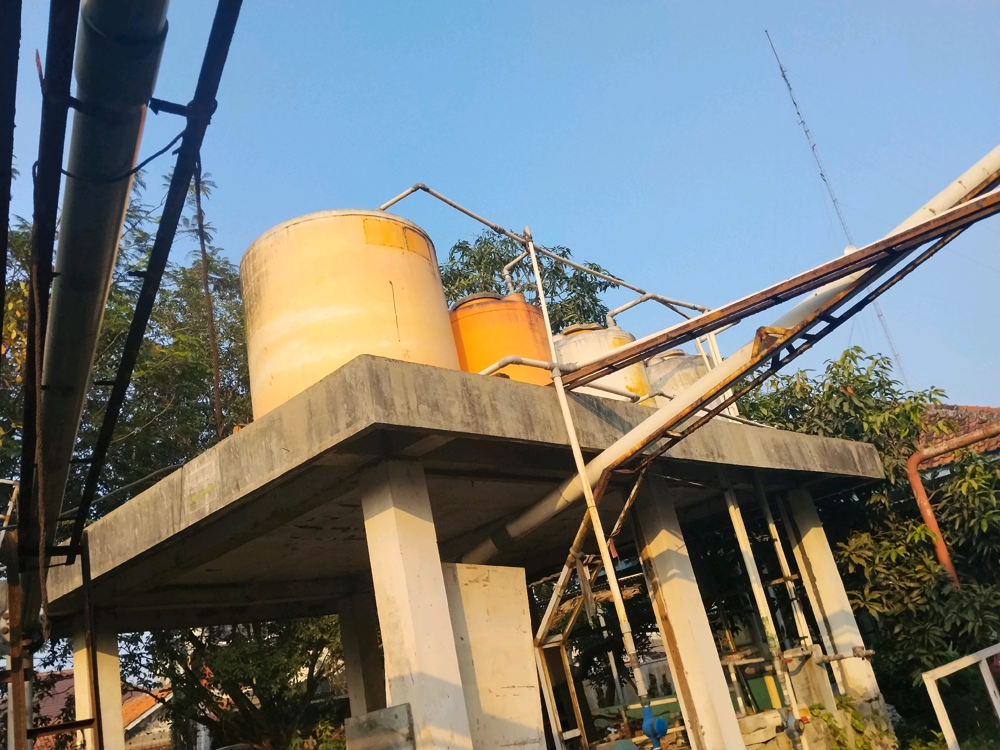 This image captures an elevated concrete platform supporting multiple water tanks. The platform is supported by several thick concrete pillars. On the platform, there are at least three cylindrical water tanks visible, one large yellow tank and two smaller orange tanks. Piping systems connect to and run between these tanks, and extend downwards from the platform, supported by metal structures. Several large pipes are also seen running horizontally and diagonally across the scene, some attached to metal framework. The background features lush green trees and a clear blue sky. In the distance, rooftops of houses are visible, suggesting a semi-urban or residential area. The overall impression is one of an industrial or utility structure integrated into a vegetated environment. The image is taken from a low angle looking upwards towards the tanks and platform. The lighting suggests it is late afternoon or early morning, with warm sunlight casting shadows. There are no people or animals visible in the frame.