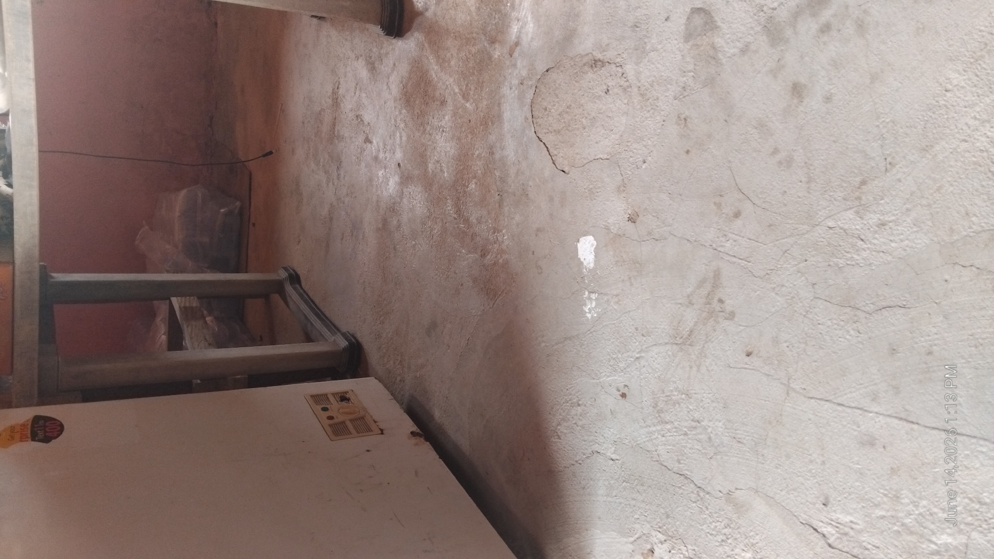 This is a close-up, ground-level, full-frame shot of a concrete floor inside a room. A white upright freezer is visible on the left, with a yellow and orange sticker on its side. Next to the freezer is a table with a grey frame and a light-colored top, with stacks of items wrapped in plastic underneath. The room appears to be in a state of disrepair, with cracks and stains on the concrete floor and a pinkish-brown wall in the background. The lighting is dim, casting shadows. The image is dated June 14, 2026, 1:13 PM. The location is identified as Howayi, Nigeria.