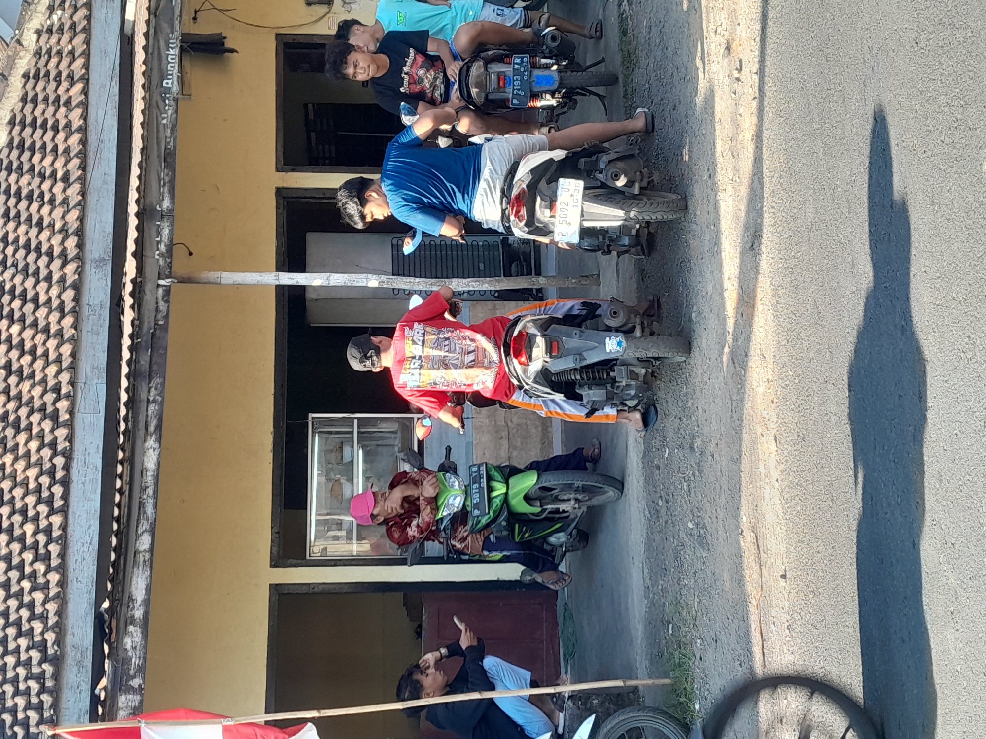 In the city of Bulurejo, Indonesia, a group of young men are gathered on motorcycles outside a building. The building has a yellow facade and a tiled roof, with several open doorways and windows. In the foreground, a person wearing a red shirt with graphics is sitting on a black and white motorcycle, facing away from the camera. To their left, another person in a blue shirt and grey shorts is also on a motorcycle, looking to the side. Further left, a person in a black jacket and light blue pants is crouching and looking at a smartphone. On the far right, a person in a black t-shirt and a person in a light blue t-shirt are standing or sitting near motorcycles. A red and white flag is visible on a pole on the far left of the image. The ground is a mix of asphalt and gravel.