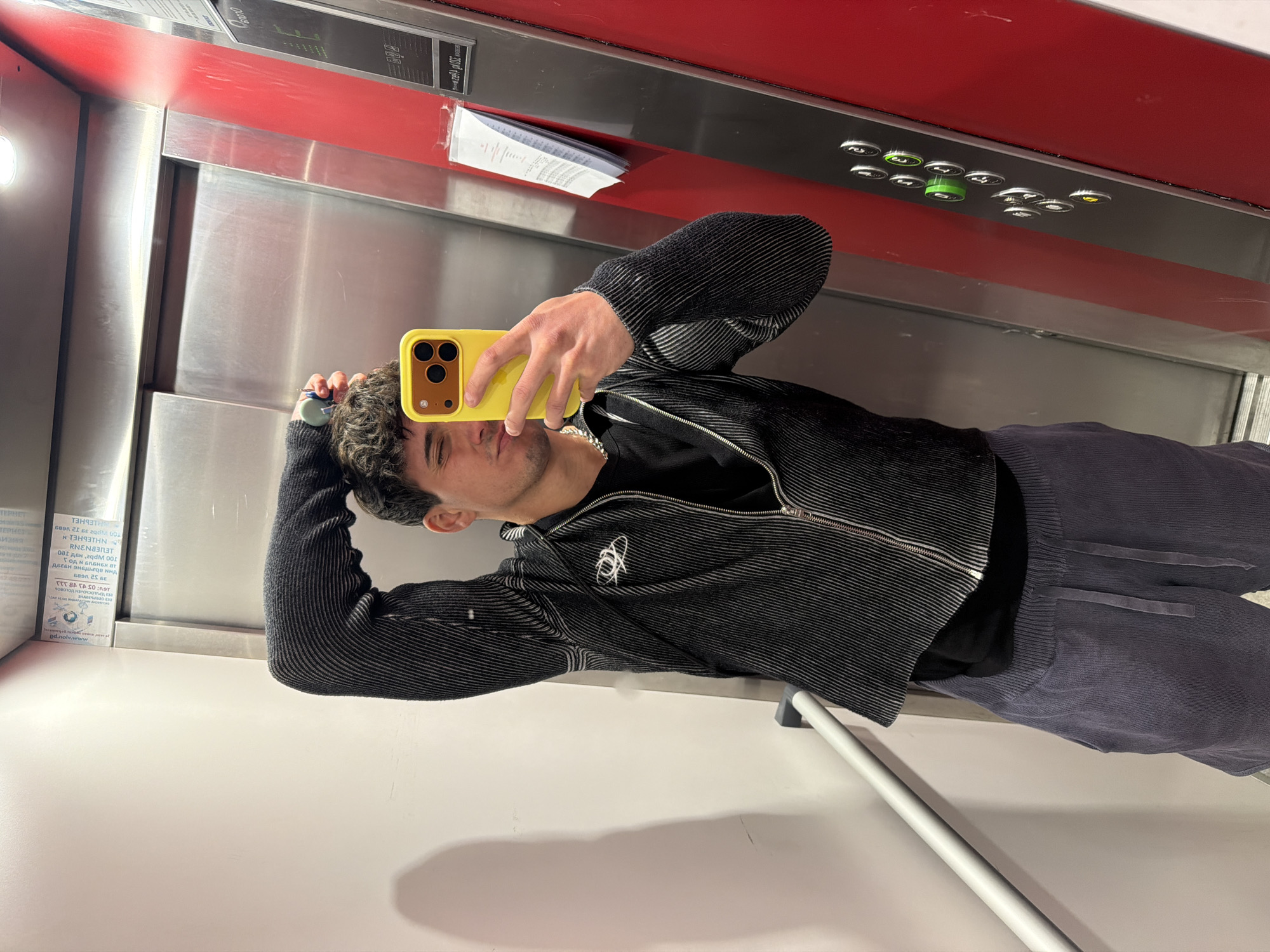 A young man takes a mirror selfie inside an elevator, captured in a portrait orientation and rotated 90 degrees counter-clockwise. He has dark, curly hair and light stubble, looking directly at his smartphone with a yellow case, which covers part of his face. He wears a black, textured, long-sleeved zippered jacket with a small white logo on the left chest, over a black t-shirt. His pants are dark gray or blue corduroy.
The setting is an elevator interior with stainless steel walls and doors, and a red panel above the door. The floor appears to be a light color. A silver handrail runs horizontally along the lower part of the wall. On the red panel, there are elevator control buttons, some of which are illuminated green, indicating active floors or selections. A white piece of paper or document is taped to the red panel above the doors, showing some mirrored text.
The visible text, which appears mirrored on the paper, includes what looks like "ИНТЕРНЕТ" (Internet) and several sequences of numbers, potentially contact information or service details, such as "002" and other numerical combinations like "39 12 1683". The OCR also picked up "100 kbps", which could relate to an internet speed detail. The overall impression is a modern, functional elevator. The image captures a casual, self-portrait moment.