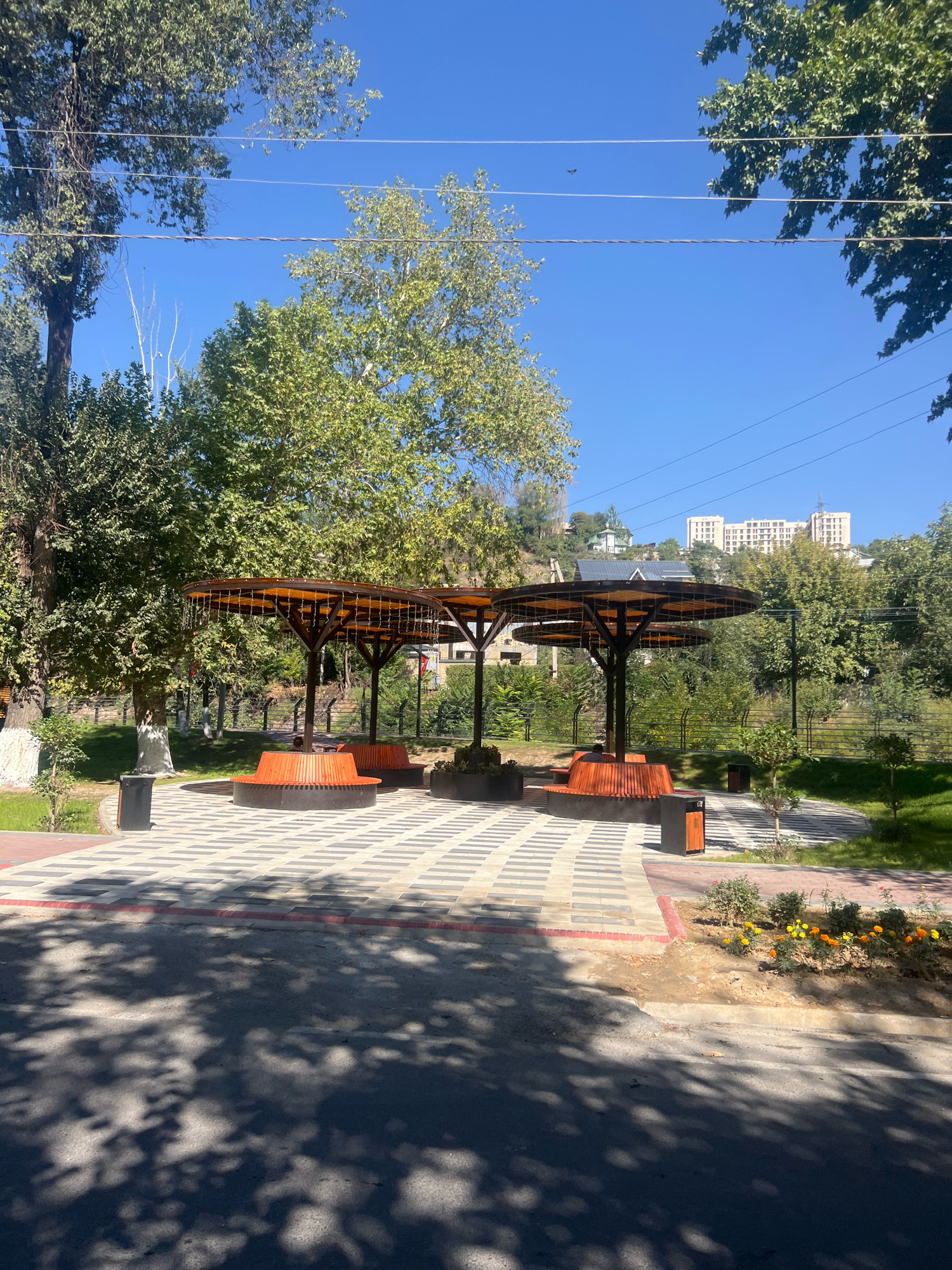 A bright, sunny day in Osh City, Kyrgyzstan is captured in this image, featuring a modern park area with distinctive circular seating and overhead shade structures. The foreground shows a paved walkway with a checkered pattern of light and dark grey tiles, bordered by a red brick trim. A portion of an asphalt road, dappled with the shadows of trees, occupies the bottom of the frame.
Dominating the mid-ground are two circular, wooden benches, each beneath a large, cantilevered, umbrella-like shade structure with an orange-tinted translucent roof. The shade structures are supported by dark, slender poles. One of the benches has a single person sitting on it, facing away from the camera. Two black trash receptacles with orange accents are positioned near the benches.
Behind the seating area, a neatly manicured lawn slopes gently upwards, bordered by a dark metal fence. Further back, trees with lush green foliage provide a natural backdrop. In the distance, on a hillside, several residential buildings and a larger, multi-story building are visible, suggesting the park is located within or near a residential area of the city. The sky is a clear, vibrant blue, with a few wispy white clouds and a small, distant bird visible. The overall impression is one of a well-maintained public space designed for relaxation and enjoyment.