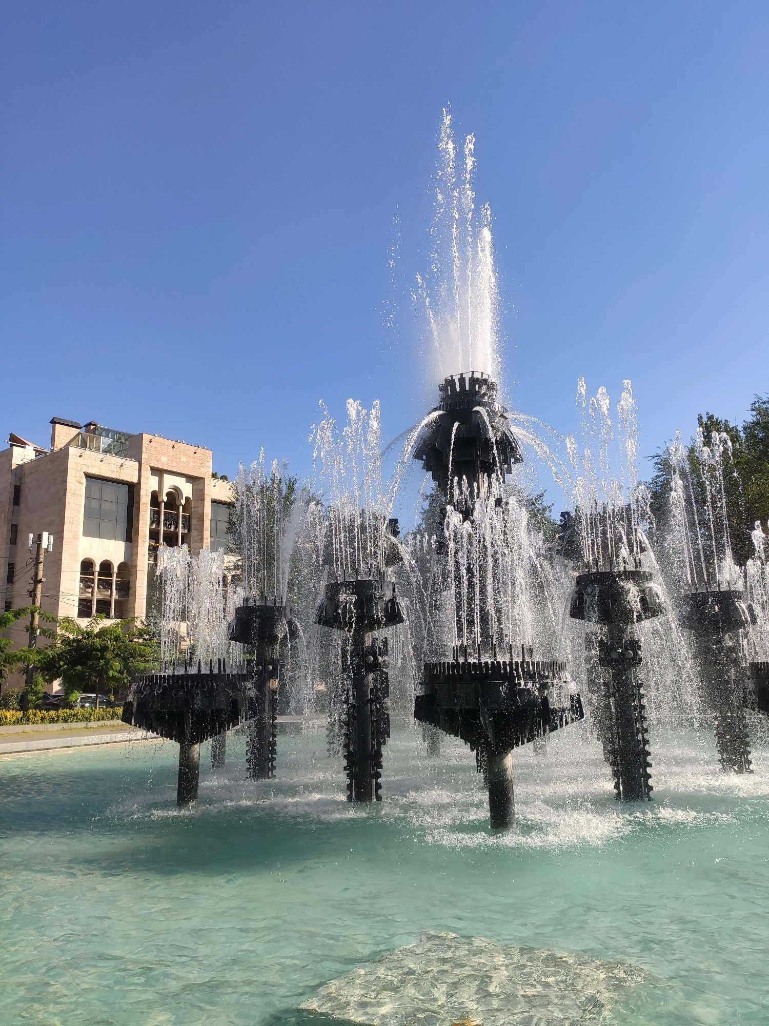 The image depicts the Cascade, a monumental stairway in Yerevan, Armenia. The photograph captures a large fountain at the base of the Cascade, with water jets spraying upwards against a clear blue sky. The fountain is intricately designed with dark, metallic structures supporting multiple tiers of water outlets. In the background, a light-colored building with arched windows and balconies, characteristic of Armenian architecture, is visible. Lush green trees and foliage frame the scene, suggesting a warm, sunny day. The water in the fountain basin is a clear, turquoise hue, reflecting the bright sunlight. No people are visible in the frame. The overall impression is one of a vibrant and well-maintained public space.