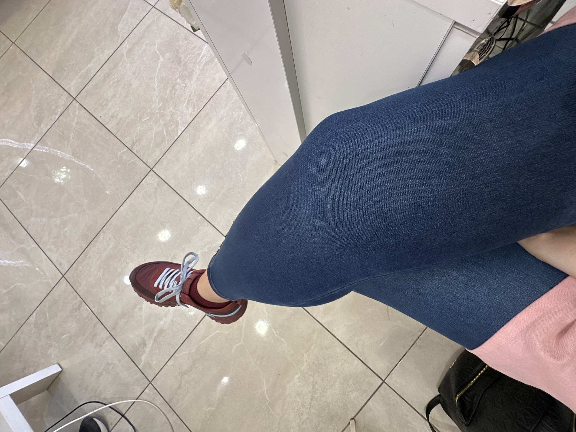 The image shows a person sitting in what appears to be a brightly lit commercial space, possibly a store or office, judging by the tiled floor and white shelving units. The person is wearing blue jeans, a pink shirt, and burgundy sneakers with white laces. A black bag is positioned near the person. The setting could potentially be in Sofia, Bulgaria, given the presence of commercial infrastructure, but there are no specific elements in the picture to confirm the location without a doubt.