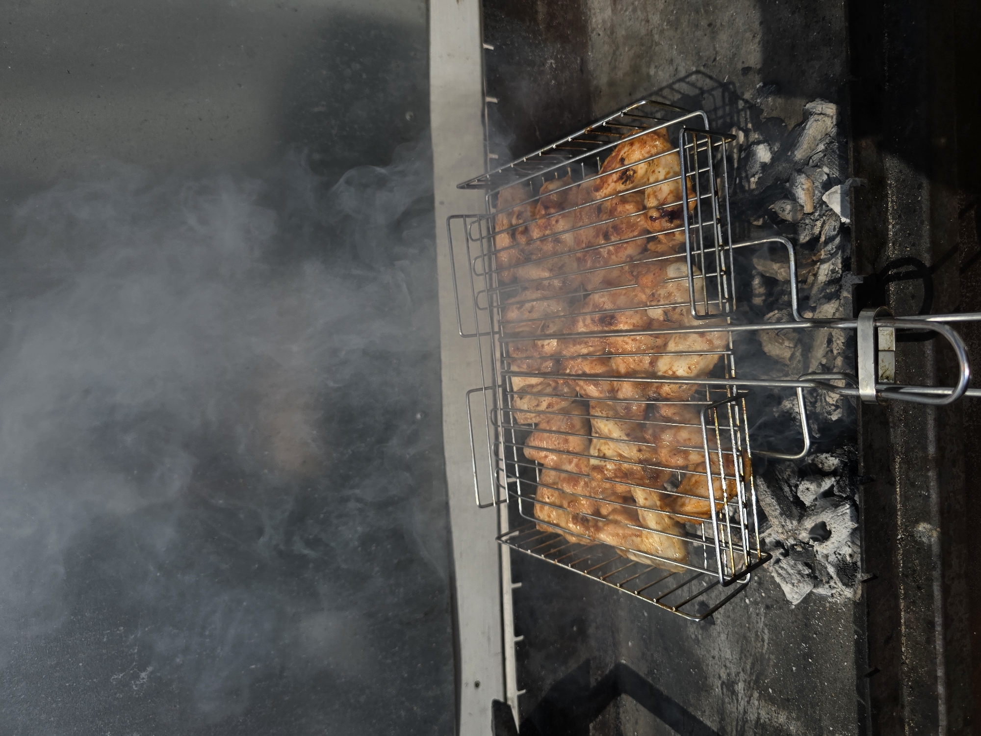 The image shows a close-up, high-angle shot of chicken legs cooking on a metal grill basket over hot coals. The chicken is seasoned and appears to be browning. Smoke is rising from the grill and obscuring the background, which appears to be a metallic or glass surface. The grill itself is placed on a dark, gritty surface, likely a barbecue pit. There are no people visible in the image. This scene is set in a backyard or outdoor cooking area, likely in Yevpatoriya, Ukraine, based on the provided context.