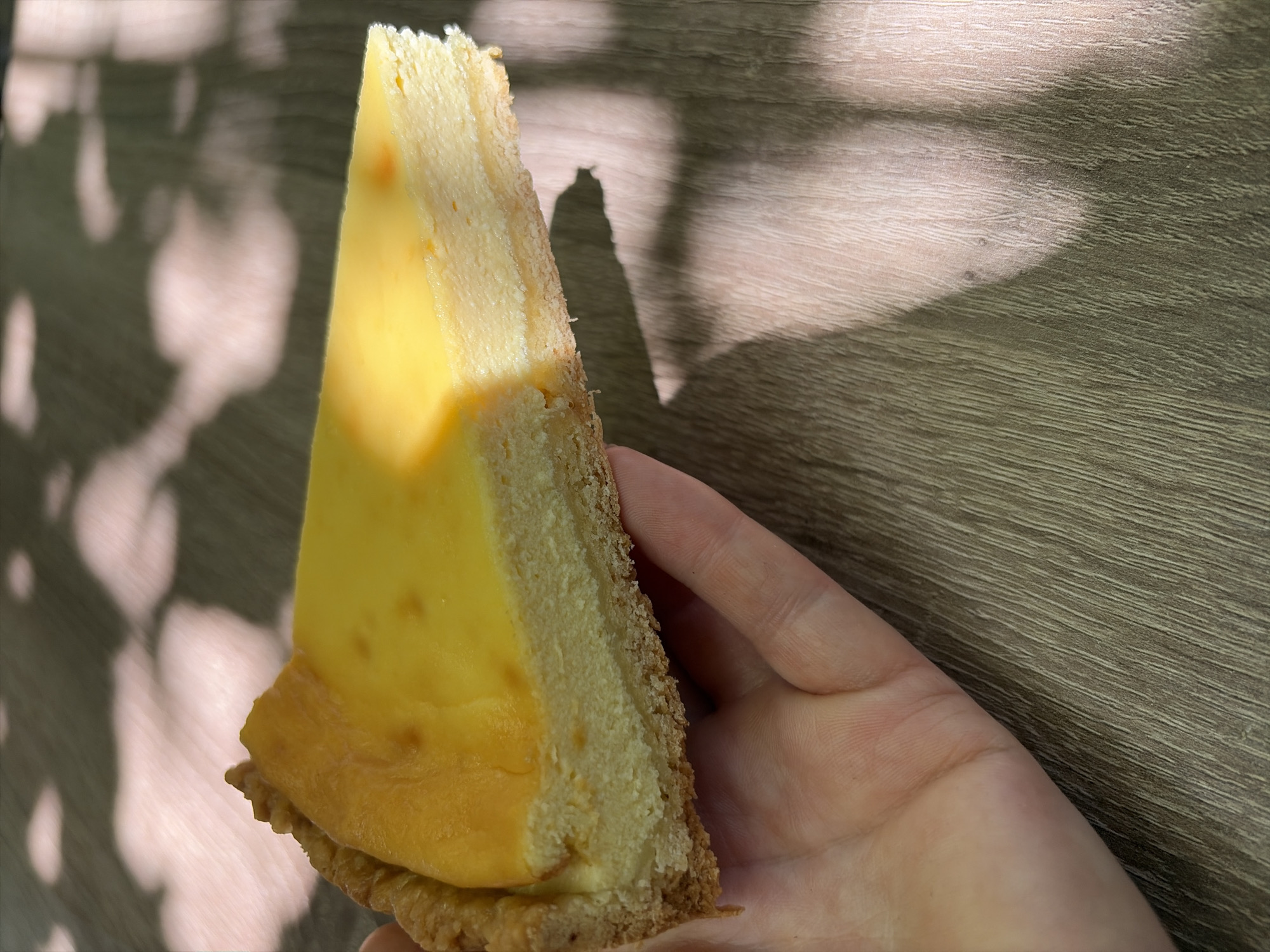 A slice of lemon tart is held in a person's hand over a wooden table. The tart has a golden-brown crust and a smooth, bright yellow filling. Sunlight casts dappled shadows on the table. The image appears to be taken indoors.