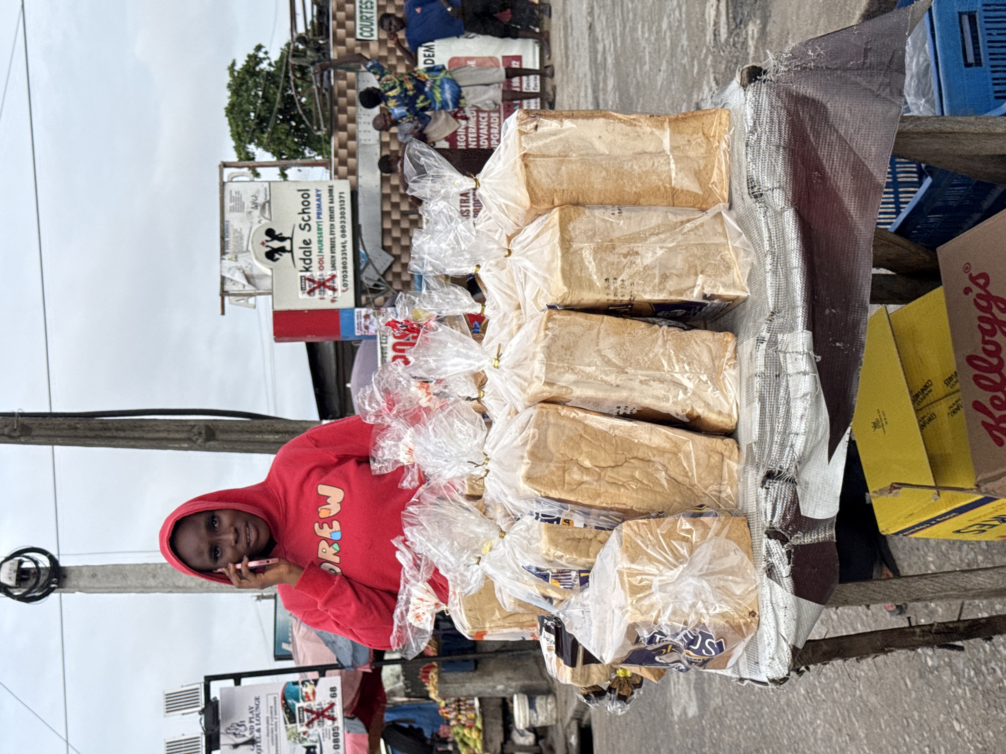A woman wearing a red hoodie, emblazoned with the word "OREW" in colorful letters, stands next to a table laden with wrapped loaves of bread in Ado, Nigeria. She is holding a pink smartphone to her ear. The bread is arranged in rows, partially covered by a metallic fabric. A Kellogg's cereal box sits near the table's edge.
In the background, street signs are visible. One sign reads "Grand Play Hotel & Lounge" with a partial phone number, and another sign indicates "Kdale School, a Goli Nursery Primary." The full address is 91A Logun Street, Even Estate, Badore, along with phone numbers 07038033141 and 08033031371. A sign behind the bread loaves includes the words "begin, intermed, advance, upgrade." People are standing near this sign in the background. A power pole is visible on the left. The sky appears overcast.