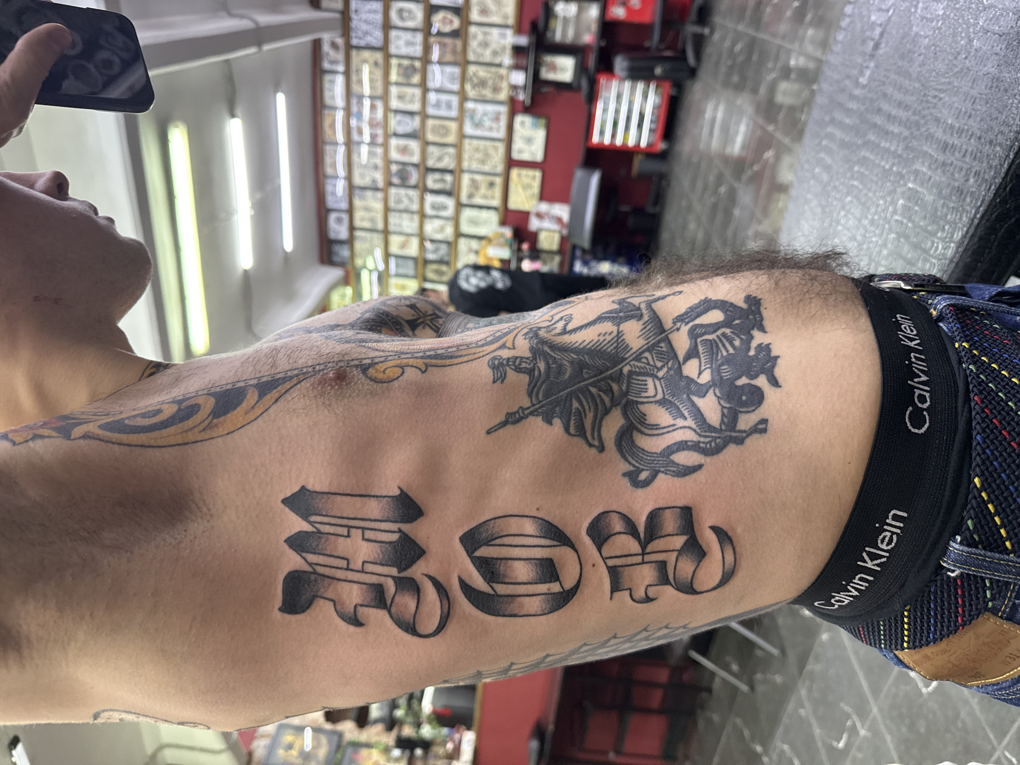 A close-up, low-angle shot depicts a person's torso, showcasing several tattoos. The tattoos include stylized letters "ROM" and an image of a figure with a spear and what appears to be a winged creature. A black Calvin Klein underwear waistband is visible, as are the top portion of patterned denim pants. The person is holding a smartphone and seems to be taking a picture of themselves. In the background, there are fluorescent lights on the ceiling, a wall covered with framed images and a partially visible red wall with a metal tool organizer. Based on the items and decor, the location appears to be a tattoo parlor. The city cannot be determined from this image.
