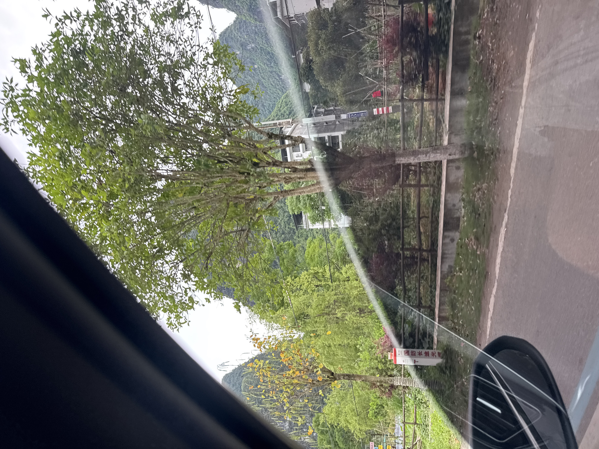 The image, taken from inside a vehicle, shows a roadside view in Guilin, China. Lush green foliage and trees line a narrow asphalt road, which runs alongside a wooden fence. In the background, there are karst mountains, a common feature of the Guilin landscape. Buildings are sparsely visible among the trees. There are sign posts with Chinese characters visible along the side of the road. The vehicle's side mirror is visible in the lower right corner. A portion of the vehicle’s interior frame is seen along the left side of the image.
