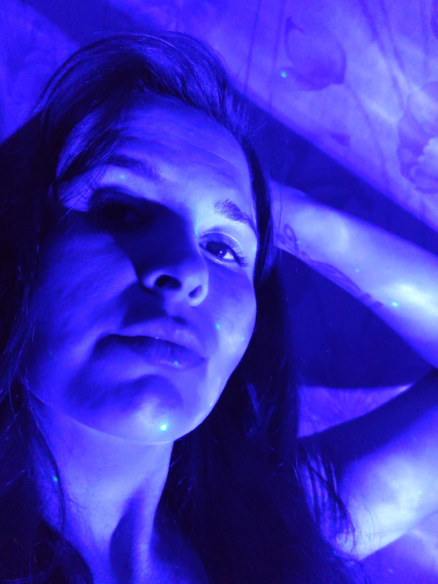 A close-up, low-angle photograph depicts a woman with dark hair illuminated by a blue light source. The woman's face, neck, and part of her upper arm are visible. Several small, green reflective points are scattered across her skin and in the background. A patterned object, possibly fabric or a wall covering, is partially visible in the upper right of the frame. The location of Фоки, Russia, is not discernible from the image.
