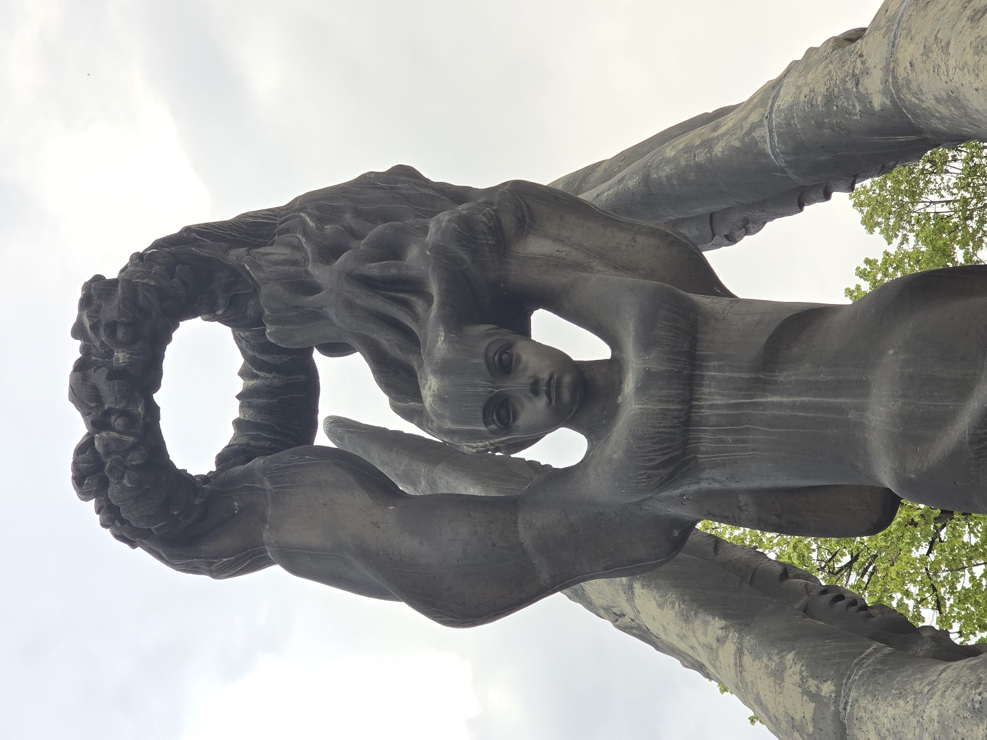 A large, dark-colored monumental sculpture, likely made of bronze or weathered stone, dominates the frame, captured from a low angle against a bright, overcast sky. The sculpture is rotated, making it appear horizontally oriented in the image. The central element is a serene female figure with a contemplative or solemn expression and long, flowing hair that blends into the sculpture's organic forms. Her eyes are large and gaze slightly downwards.
The figure is integrated into a larger, sweeping composition. To her left, she is connected to or emerges from a large, textured, circular or wreath-like element. To her right, the sculpture extends into broad, curved, wing-like or abstract shapes. The material has a rough, aged texture, with visible lines and variations in color that suggest weathering.
In the background, visible through apertures in the sculpture's structure, are bright green leaves of trees, indicating an outdoor setting such as a park or public square. The sky is uniformly cloudy, suggesting an overcast daytime. A very small airplane is faintly visible high in the sky in the upper left background. No text is visible on the sculpture or in the surrounding environment. The artwork conveys a sense of solemnity and artistic majesty.