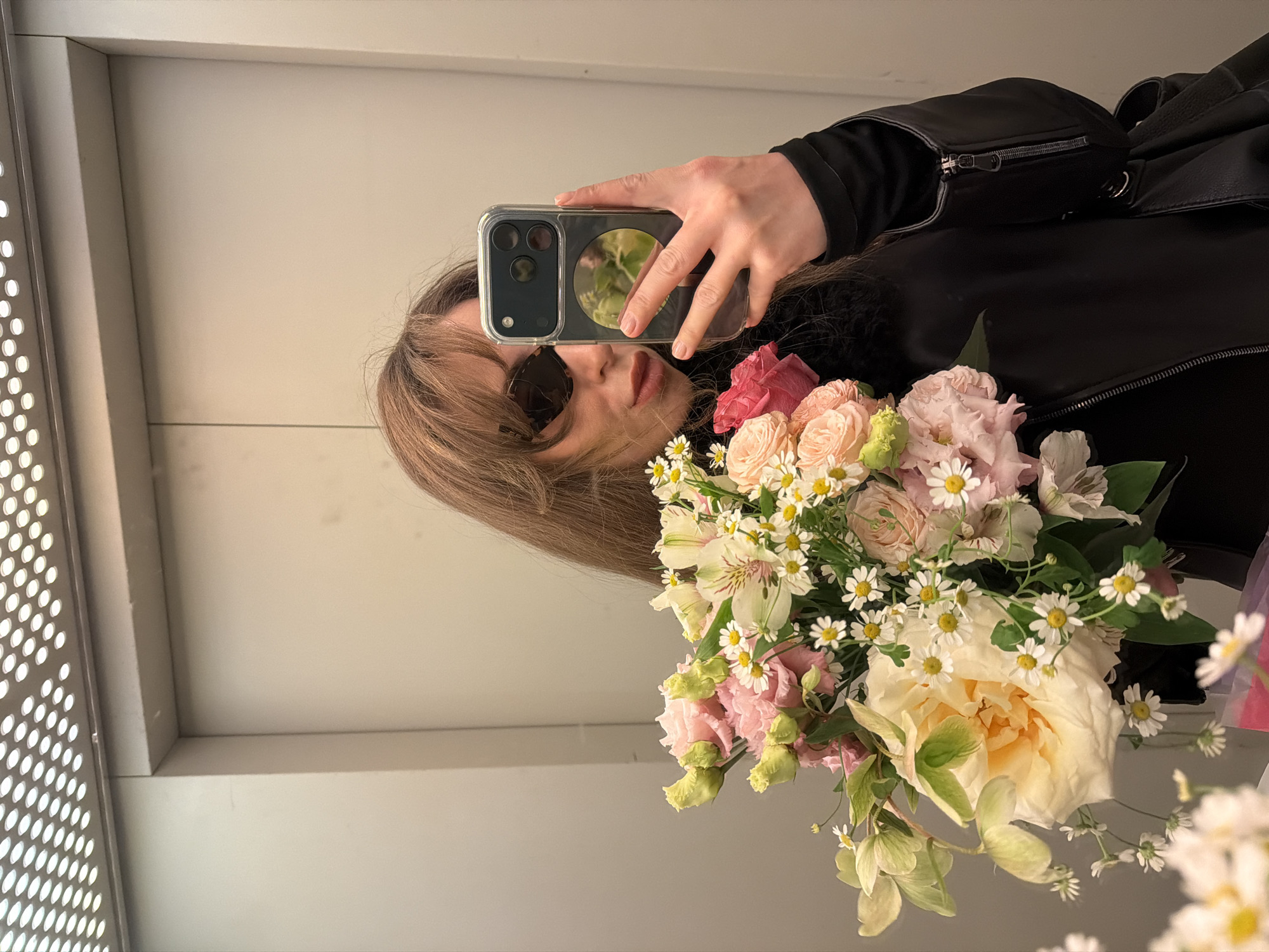 A female individual takes a mirror selfie inside an elevator in Moscow, Russia. The subject, with long brown hair, wears black cat-eye sunglasses, a black long-sleeved top or jacket, and muted red/brown lipstick. She holds a dark-colored smartphone in a clear case with her right hand, angled to capture her reflection. A large, vibrant bouquet of fresh flowers is held in front of her, obscuring part of her body. The bouquet prominently features cream and pink roses, pink lisianthus buds, white and yellow daisies, and green leafy accents. The elevator interior has plain, light-colored wall panels and a metallic ceiling panel with numerous circular light fixtures.
