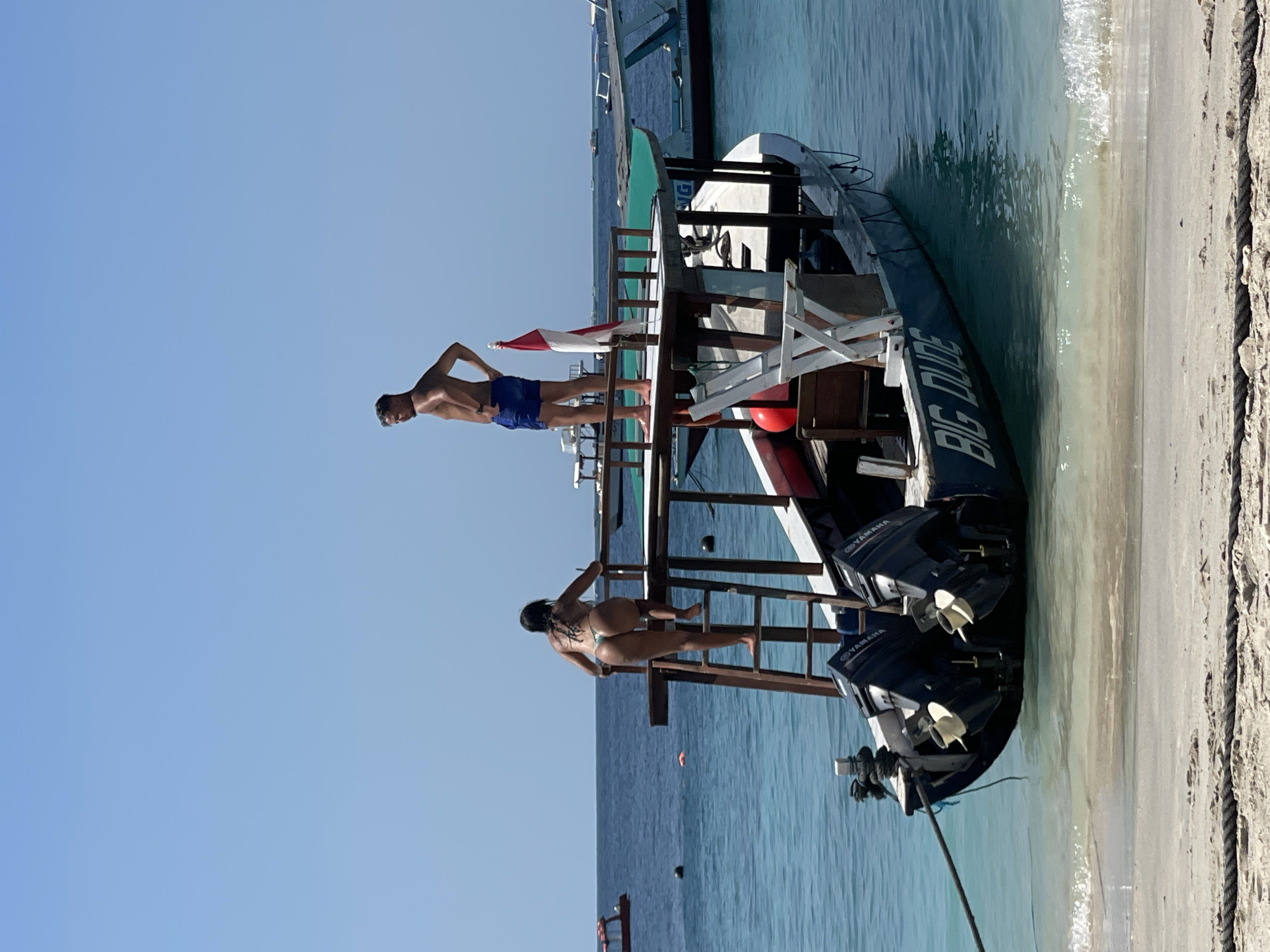 An adult male wearing blue swim trunks stands with his hands on his hips on the upper deck of a boat labeled "BIG DUDE" which is moored in shallow, clear blue water near a sandy beach. Next to him, an adult female wearing a patterned bikini climbs a wooden ladder attached to the side of the boat, ascending towards the upper deck. The boat has two outboard motors at the stern. The sky is clear and blue, and the sun is shining.