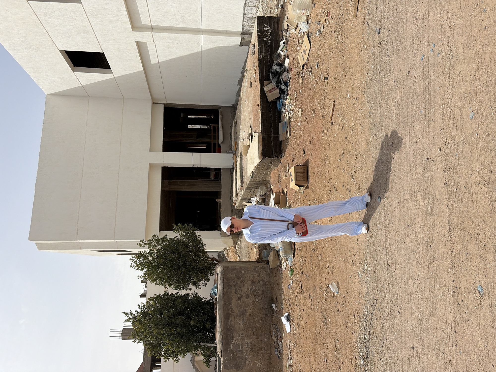 A man in a white hat, sunglasses, and a light blue striped outfit stands on a sandy area next to an unfinished white building in Sharm El Sheikh, Egypt. Debris and litter scatter the ground. Trees are visible near the building. His shadow stretches out across the ground.