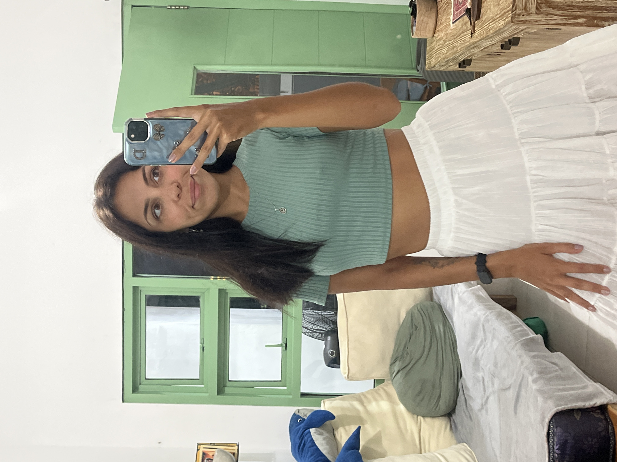 The image, originally rotated 90 degrees counter-clockwise, captures a young woman taking a selfie indoors in Kuta Selatan, Indonesia. She stands slightly to the left of the frame, holding a light blue/grey smartphone encased in a clear cover adorned with a butterfly-like charm and a prominent letter 'D'.
The woman has long, dark hair, dark eyes, and a subtle, pleasant smile as she looks slightly upwards and to her left. She is dressed in a light green, ribbed crop top and a flowing, tiered white skirt. A delicate silver necklace with a small, circular pendant featuring an infinity symbol or stylized '8' is visible around her neck, and she wears a dark-colored smartwatch on her left wrist.
The background reveals a brightly lit interior space with white walls. Two light green-framed windows are prominent, suggesting daytime and allowing natural light into the room. To the right, a section of wooden furniture, possibly a table or shelf, holds woven baskets and other decorative items. Below this, a light-colored sofa or daybed is visible, furnished with cream cushions and a sage green pillow, alongside a blue shark plush toy. A dark, circular object, potentially a fan or speaker, is partially visible behind the sofa. The scene conveys a relaxed and casual atmosphere.
