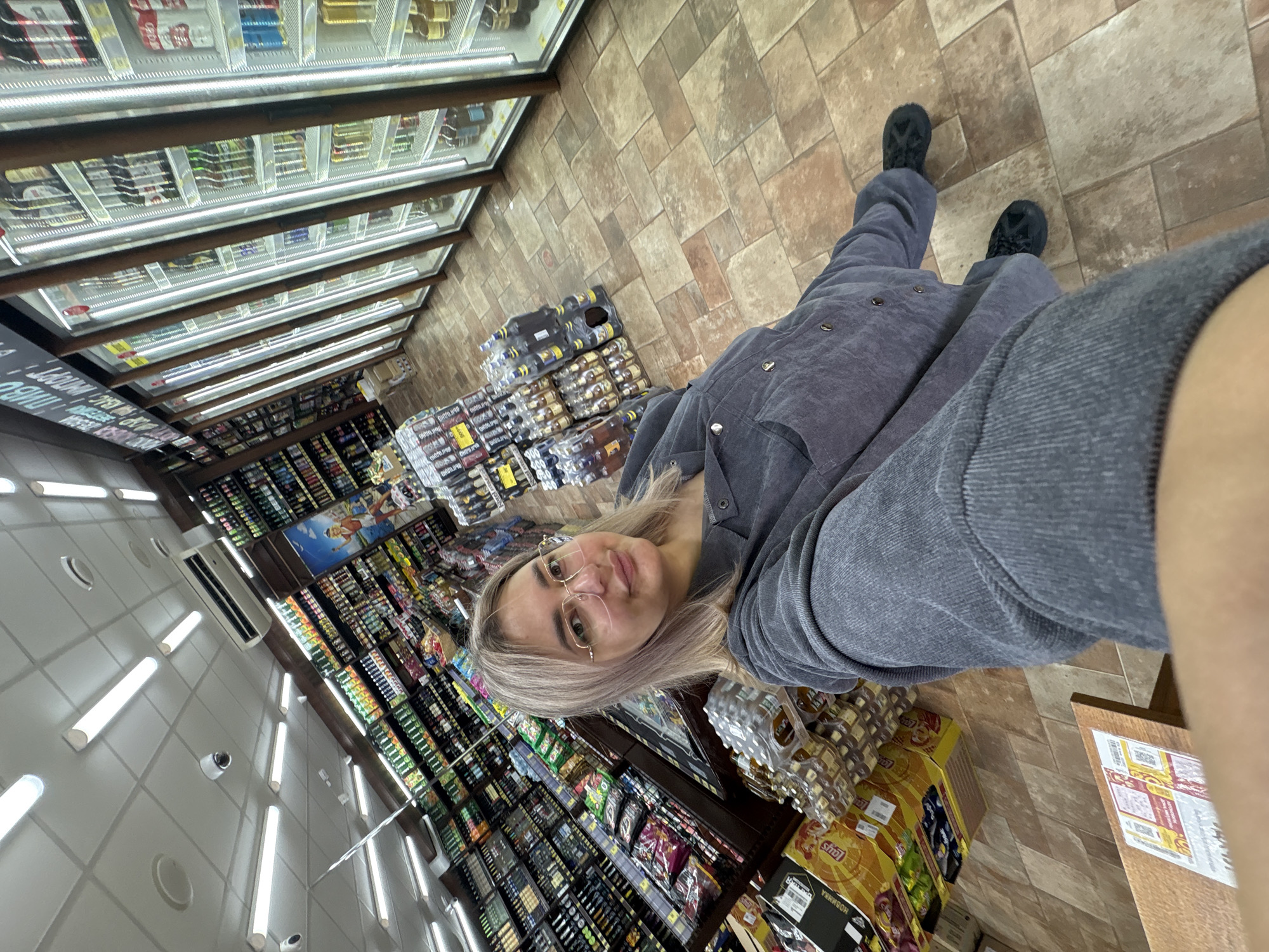 A woman with blonde hair and glasses stands in a grocery store aisle, taking a selfie. She is wearing a gray corduroy outfit and black shoes. The aisle is filled with shelves stocked with drinks and snacks. In the background, a row of glass-fronted refrigerators display more beverages. The floor is tiled with large, square tiles. The scene appears to be a typical retail environment, with lighting fixtures and security cameras visible on the ceiling. The overall impression is one of everyday commerce in a well-lit store.