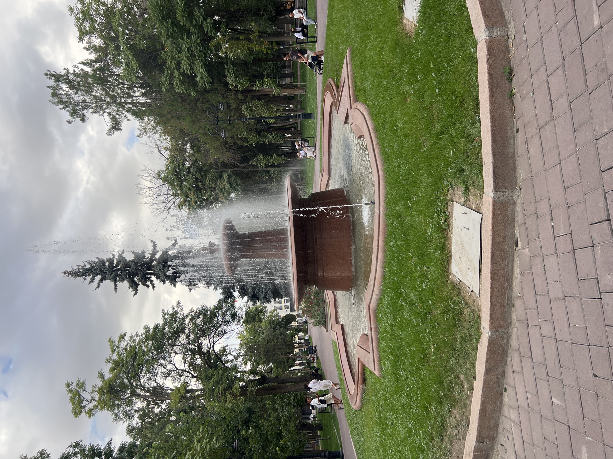 The image shows a park with a large red stone fountain in the center, spraying water upwards. The fountain is surrounded by a shallow pool and then a circular area of green grass. A paved walkway, made of red brick pavers, borders the grass in the foreground. In the background, there are many trees, some of them tall and slender, others more broad. A few park benches are visible along the walkway, with several people seated on them. Two young women in white outfits are walking on the path to the left of the fountain. The sky is overcast with clouds. The scene appears to be in an urban park, likely in Bishkek, Kyrgyzstan, given the presence of trees and typical park landscaping.