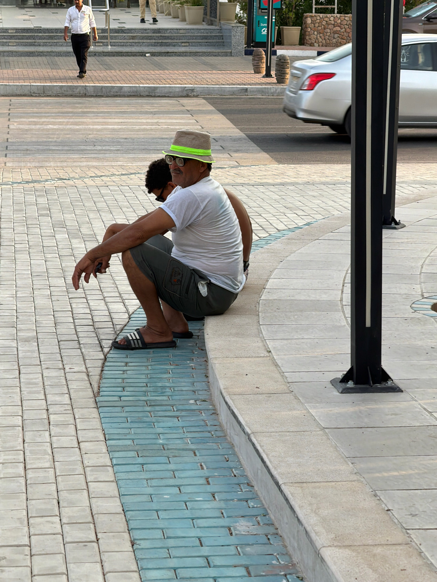 A man wearing a white t-shirt, khaki shorts, a light-colored fedora hat with a bright green band, and dark flip-flops is sitting on the edge of a sidewalk. He is resting his arms on his knees, and his head is turned slightly to his left. His glasses are perched on his nose. Another person, whose head and shoulders are visible behind the seated man, is wearing sunglasses. The ground is paved with light-colored and blue rectangular bricks. A silver car is driving on the road in the background. The scene appears to be in a public outdoor area, likely a promenade or pedestrian walkway, consistent with locations in Sharm El Sheikh, Egypt, known for its coastal resorts and leisure areas.