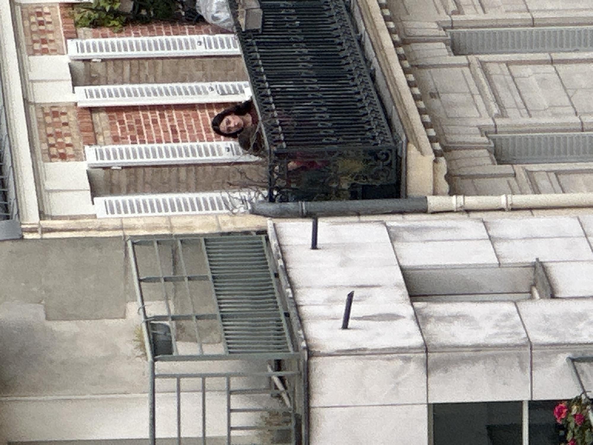 This high-angle urban photograph captures a person leaning from a balcony amidst a backdrop of contrasting building architectures. The perspective is from above, looking down onto the rooftops and facades of several structures.
In the upper-middle section of the image, a person, likely a woman with dark hair, is visible from the chest up, peering out and slightly upwards from behind an ornate black metal balcony railing. Her expression appears neutral, suggesting observation. The building she is on features a facade of patterned brickwork, with segments of reddish-brown bricks interspersed with lighter-colored, possibly stuccoed, sections. Below window openings, white, slatted grate-like structures are present. Green foliage and a white, possibly tarp-covered object are also visible on the balcony area.
To the right of this building, another structure rises, characterized by a light-colored stone or concrete facade with classical architectural details, including horizontal moldings and recessed paneling. Below the primary balcony and extending to the left, a more modern, light grey or white building section is visible. This lower structure includes flat roof areas with several dark, vertical pipes or vents. A metal grid-like structure, possibly a fire escape or industrial platform, is attached to this section. A small patch of green growth clings to the wall near this structure, and a hint of pink flowers is visible in the bottom right corner of the frame.
The scene is illuminated by diffused daylight, suggesting either an overcast sky or indirect sunlight. The architectural styles, particularly the ornate balcony and classical elements, align with typical Parisian buildings. No visible text is present in the image.