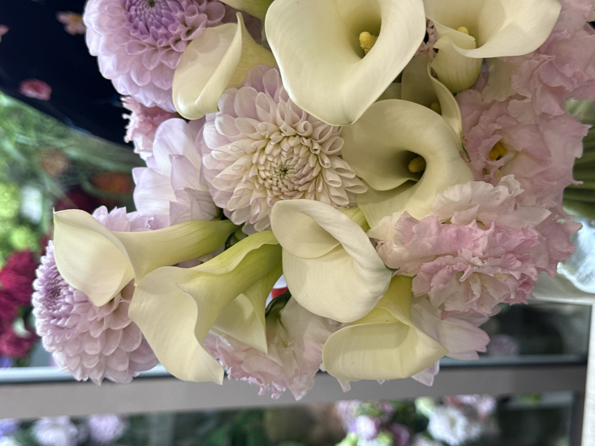 A close-up, eye-level shot showcases a floral arrangement, dominated by pale yellow calla lilies and soft pink dahlias. The background is blurred, revealing hints of a window with reflections and other out-of-focus greenery and floral elements. The image does not provide any information about the city of Assinovskaya, Russia, nor does it depict any people, actions, or interactions. It is a still-life composition of flowers.