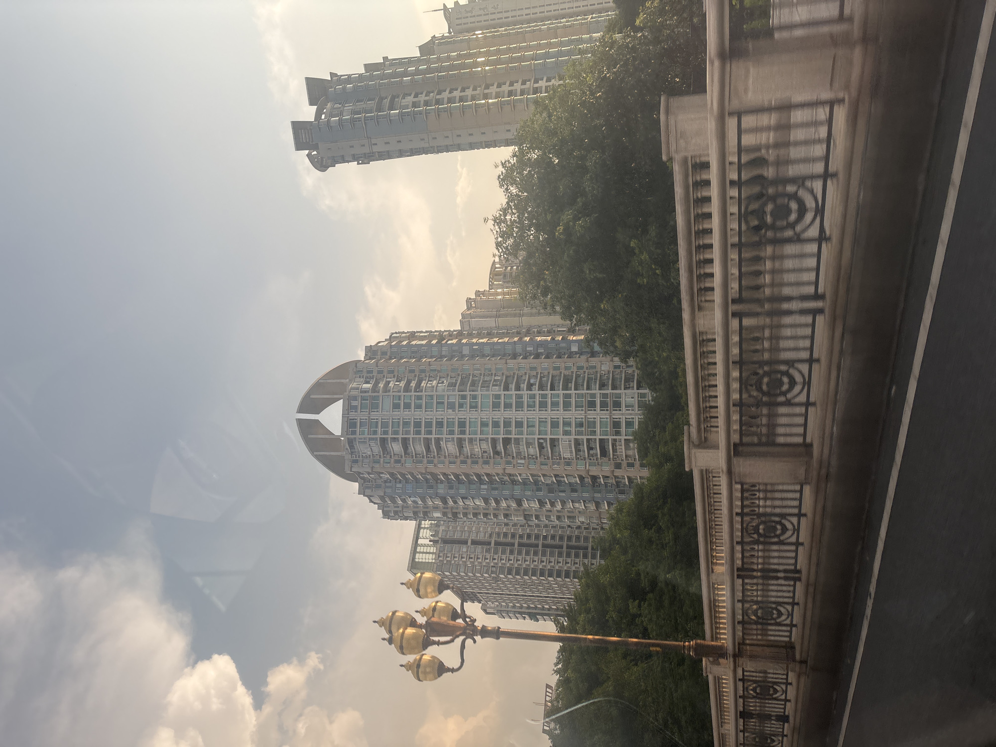 The image shows a view of modern high-rise apartment buildings in Haizhu District, China, from a street level perspective. In the foreground, a bridge railing with decorative metalwork and stone pillars is visible, suggesting the vantage point is from a moving vehicle on a road. A traditional lamppost with multiple ornate globes stands to the left of the frame. Behind the bridge, lush green trees obscure the lower portions of the buildings. The apartment buildings themselves are tall, with numerous balconies and windows, exhibiting contemporary architectural designs. The sky above is a hazy blue with scattered white clouds, indicating daytime, possibly late afternoon or early morning given the soft lighting. Reflections are visible on the glass surfaces of the buildings and potentially on the camera lens or a window through which the photo was taken. No people are clearly discernible in the image.