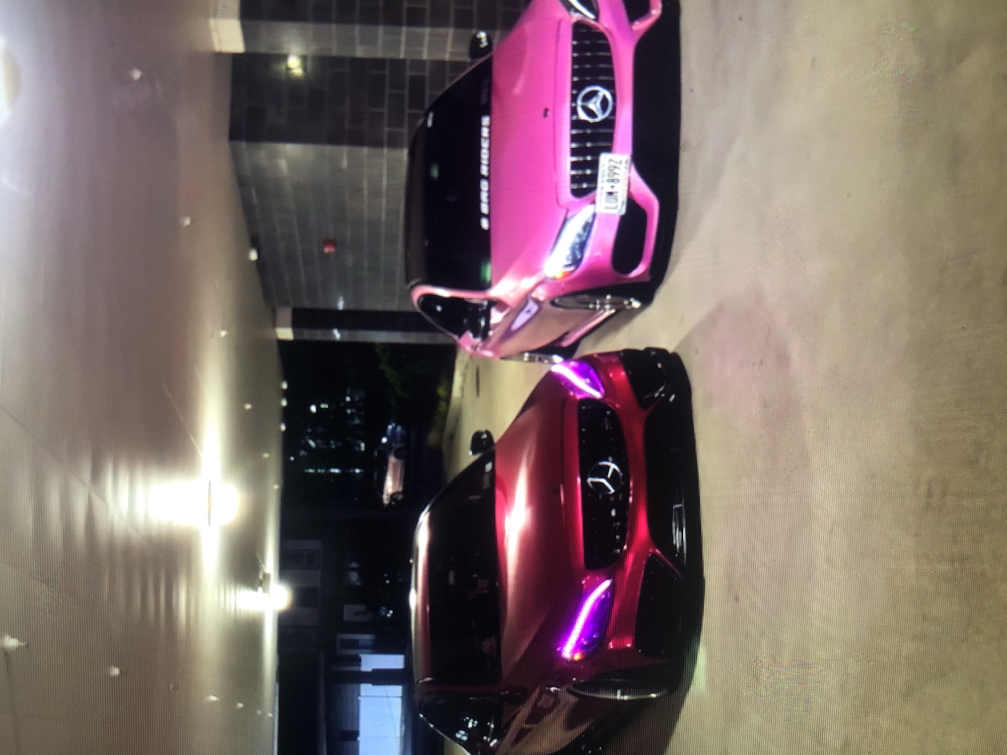 The image shows two parked cars. The first car is pink and has a license plate that appears to read "LUM-8992". "S SNG RIDERS" is visible on the windshield. The second car, which is closer, is red/maroon with purple highlights. The scene appears to be in a parking area near buildings. There is no information provided in the image about the city of Lhokseumawe, Indonesia.