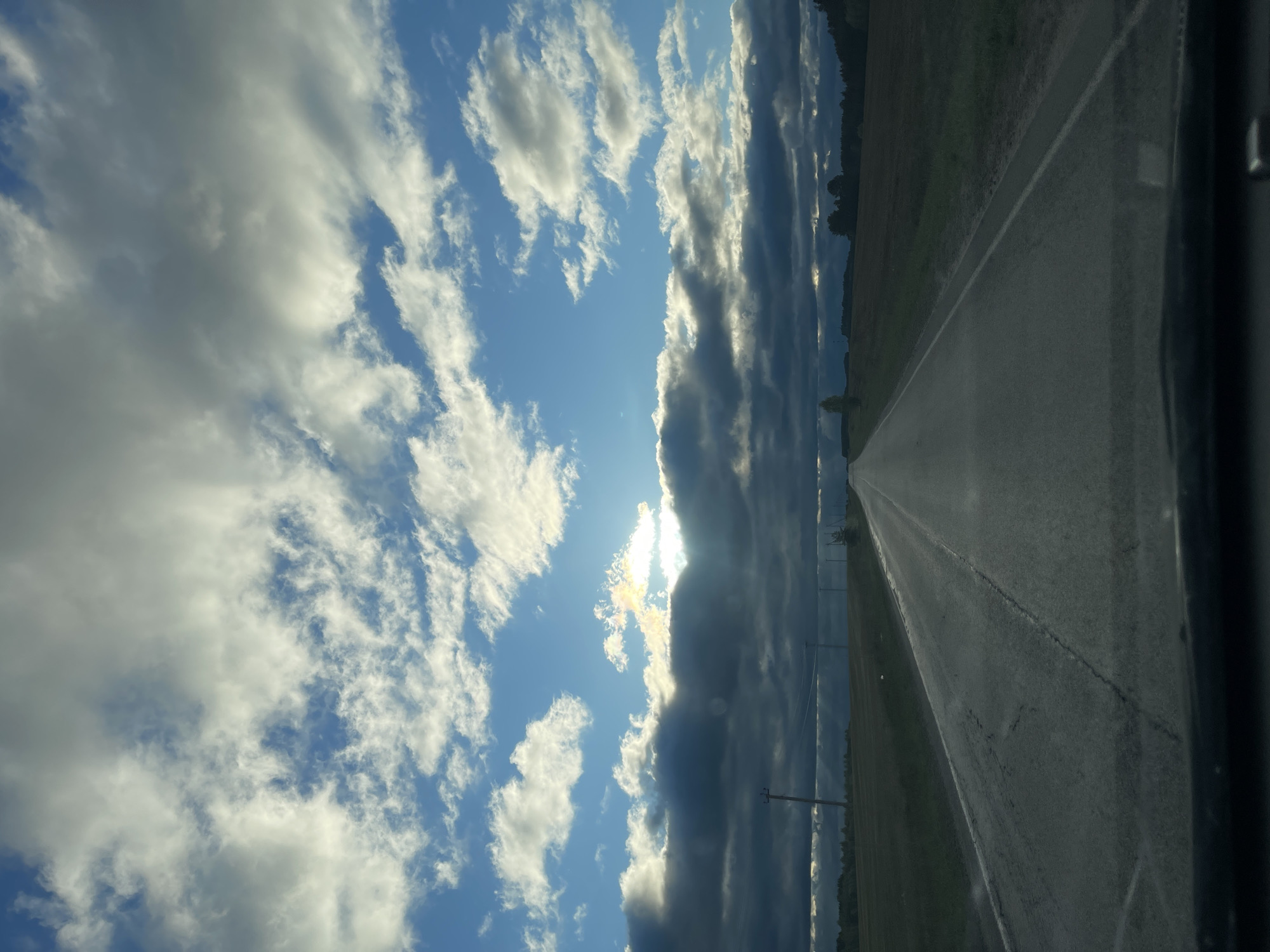 The image depicts a rural highway in Перевозное, Russia. The road stretches into the distance, flanked by fields and trees. The sky is predominantly blue with scattered white clouds, partially obscured by darker, striated clouds. The sun is visible through a break in the clouds, creating a bright glare. The image appears to be taken from inside a vehicle, with a portion of the vehicle's interior visible on the right side of the frame. There are power lines along the roadside.