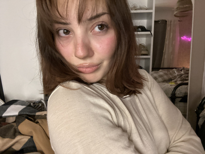 A young woman with shoulder-length brown hair and bangs poses for a selfie. She wears a light-colored, long-sleeved shirt. Her skin is fair, and she has a hint of blush on her cheeks. Behind her, a cluttered room includes a shelf with shoes, a bed with patterned bedding, and a pink neon sign. A blanket is draped in the lower left corner. The overall lighting is soft, creating a casual and intimate atmosphere.