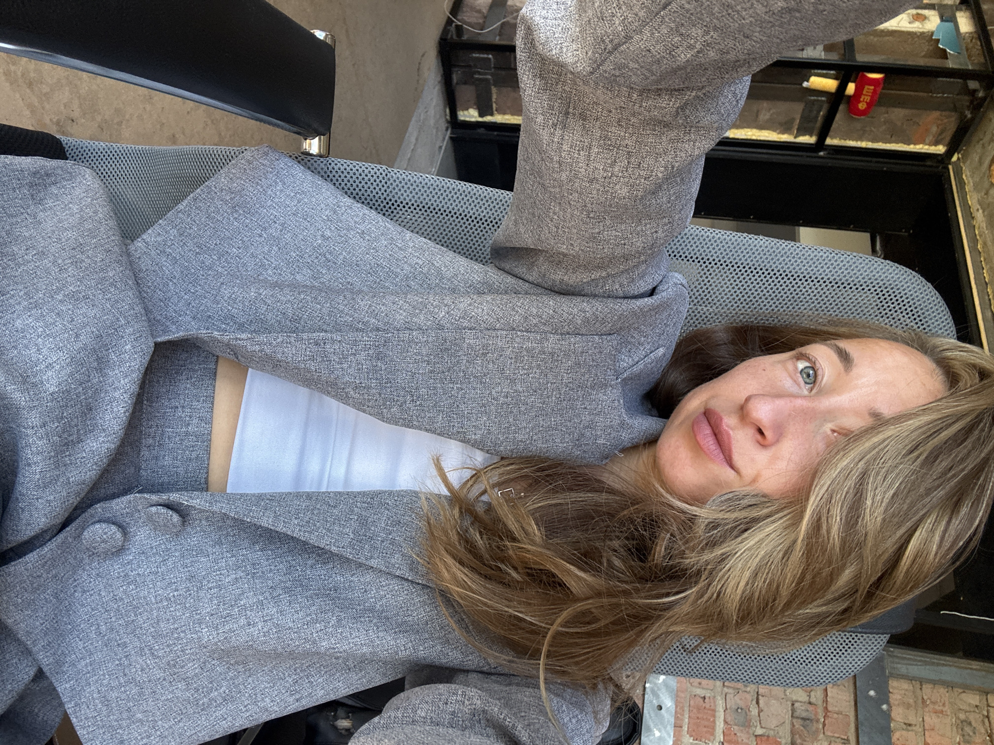 In Chaikovsky, Russia, a woman with blonde hair and green eyes, wearing a grey pantsuit and a white top, takes a selfie while sitting in a grey office chair. She is positioned in what appears to be an outdoor or semi-outdoor space with a brick wall in the background, suggesting a workshop or industrial area. Sunlight illuminates her face. To the left of the frame, a red and yellow object resembling a hammer with the Cyrillic letters "ШЕФ" is visible on a wall. The environment suggests a functional or work-related setting.