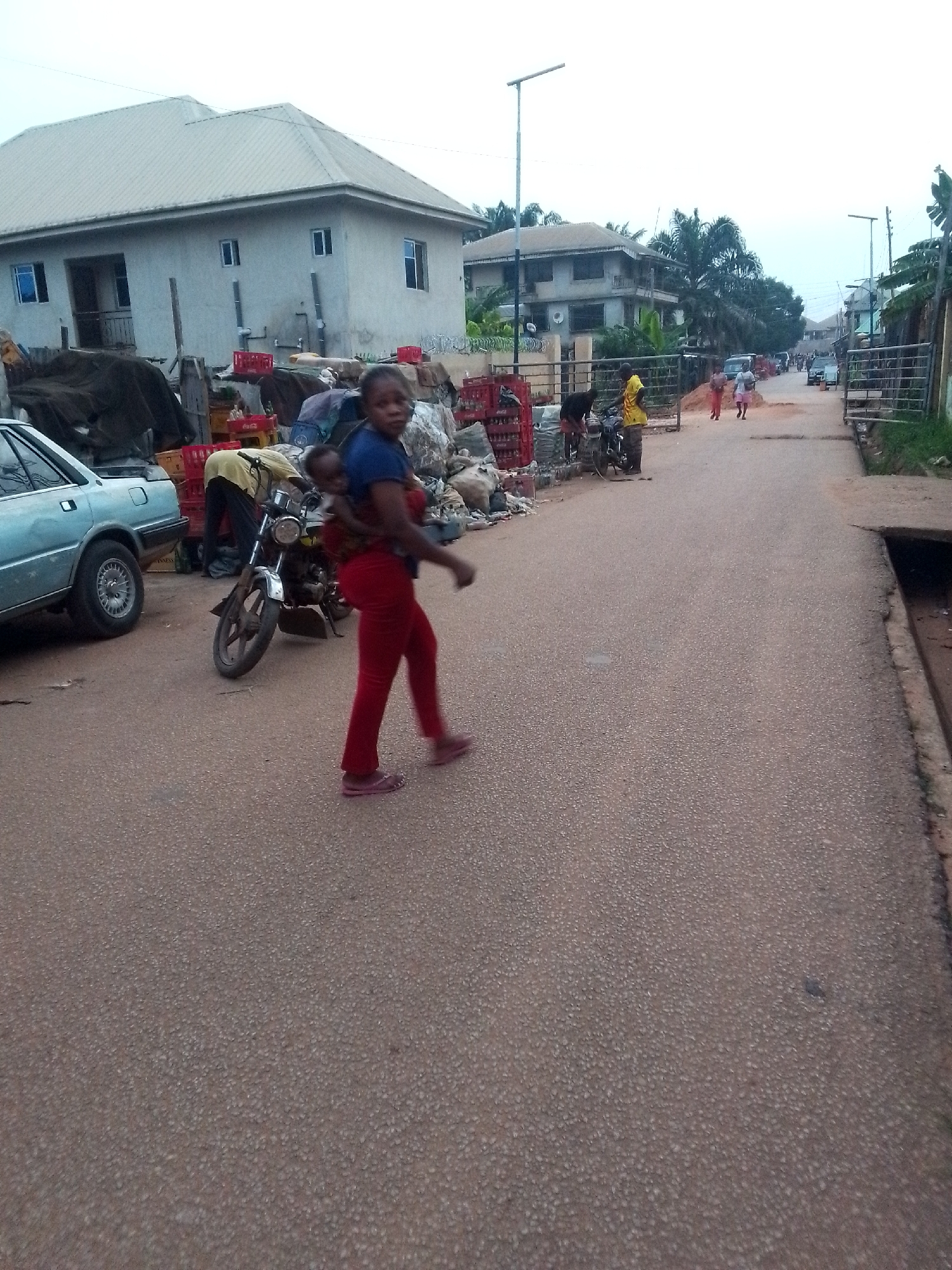The image captures a vibrant street scene in Orlu, Nigeria, under an overcast sky during what appears to be daytime.
In the foreground, a woman in a blue top and red pants walks left-to-right across the paved street, carrying a child securely on her back. She wears pink sandals. To her left, a light blue car is parked partially on the roadside. Next to the car, a man in a yellow shirt is bent over a motorcycle, possibly repairing it, with various items and covered goods piled behind him. Numerous red crates, some clearly branded "Coca-Cola," are stacked along the side of the road, suggesting a commercial or storage area. Behind these roadside goods, a light-colored, two-story building with a light-toned roof is visible.
Further down the street, more people can be seen walking, and several other buildings with varying architectural styles are present. Tall streetlights line the road, and lush green foliage, including palm trees, indicates a tropical environment. The road itself is paved with a granular texture. The overall scene depicts a typical daily activity in a bustling street environment. No strong emotions are overtly displayed, rather a sense of everyday life and movement.