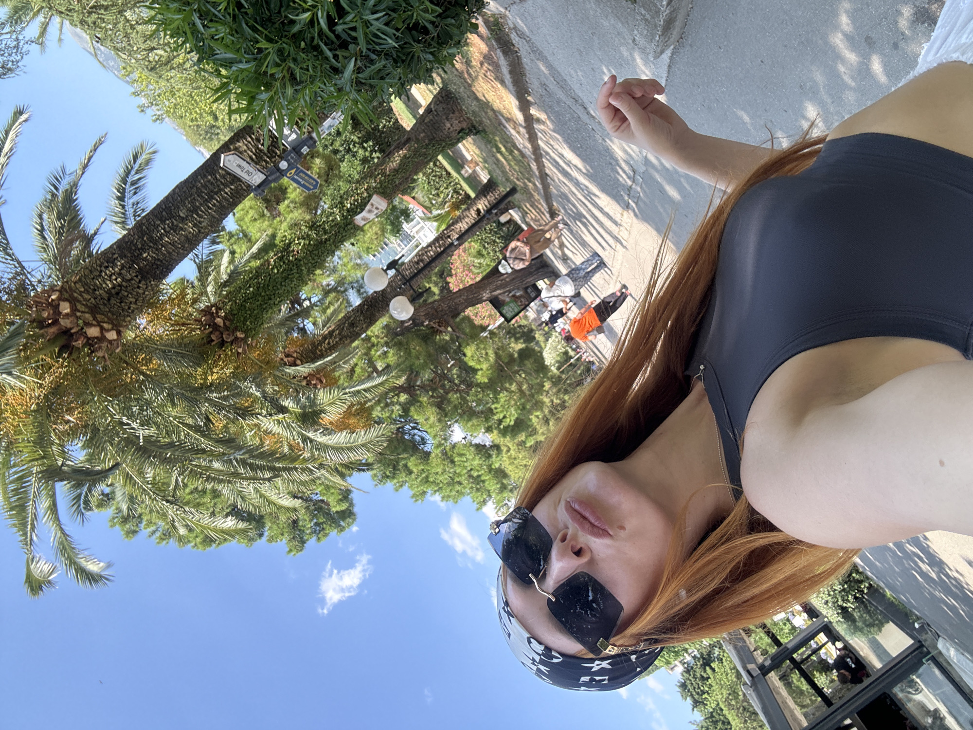 A woman with long, auburn hair is taking a selfie outdoors. She is wearing large, dark sunglasses and a black bandana with a white pattern. Her top is black and sleeveless. The background features palm trees, lush green foliage, and a bright blue sky with a few scattered clouds. In the distance, several people are walking along a paved pathway. A signpost with directional arrows is visible on a palm tree trunk, one arrow pointing to "Old Town." The scene appears to be in a park or along a promenade in Budva, Montenegro. The lighting suggests it is daytime, likely sunny.