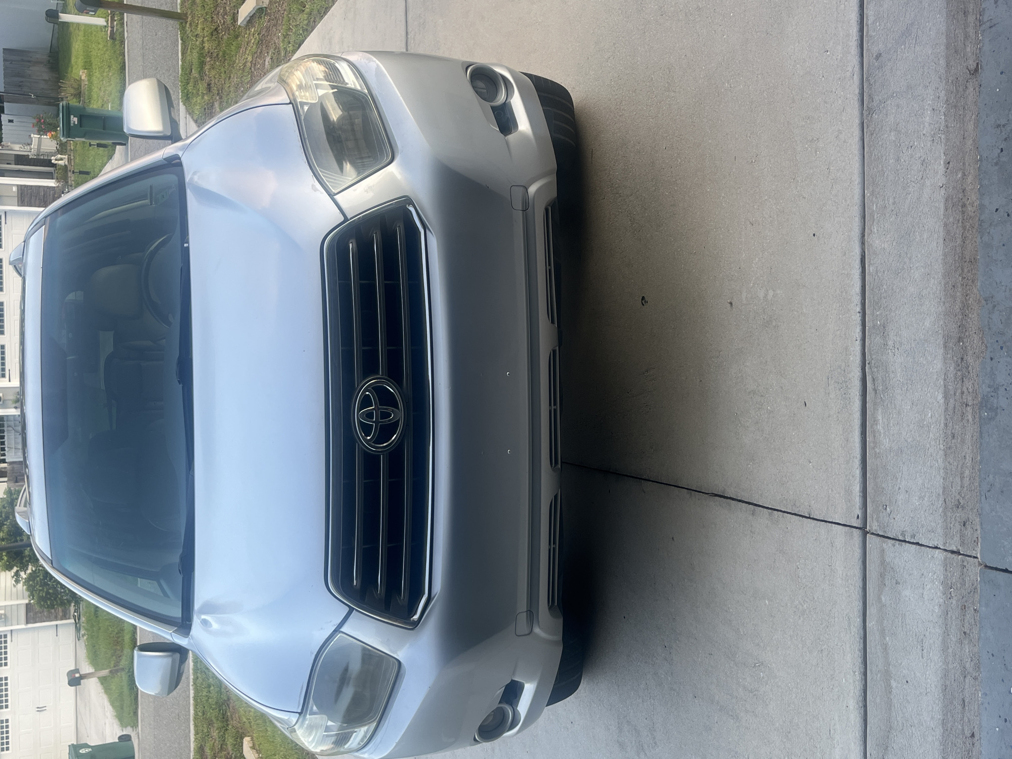 A silver Toyota Highlander SUV is parked on a concrete driveway in a residential area, likely Jacksonville, United States. The SUV is facing the camera, and its front end, including the grille, headlights, and bumper, is clearly visible. The vehicle appears to be in good condition, with no visible damage or defects. The driveway is clean and well-maintained, with some cracks and expansion joints visible. In the background, a suburban house with a white garage door can be seen, along with some greenery and a green trash can. The lighting suggests it is daytime, possibly late afternoon or early morning due to the soft shadows.