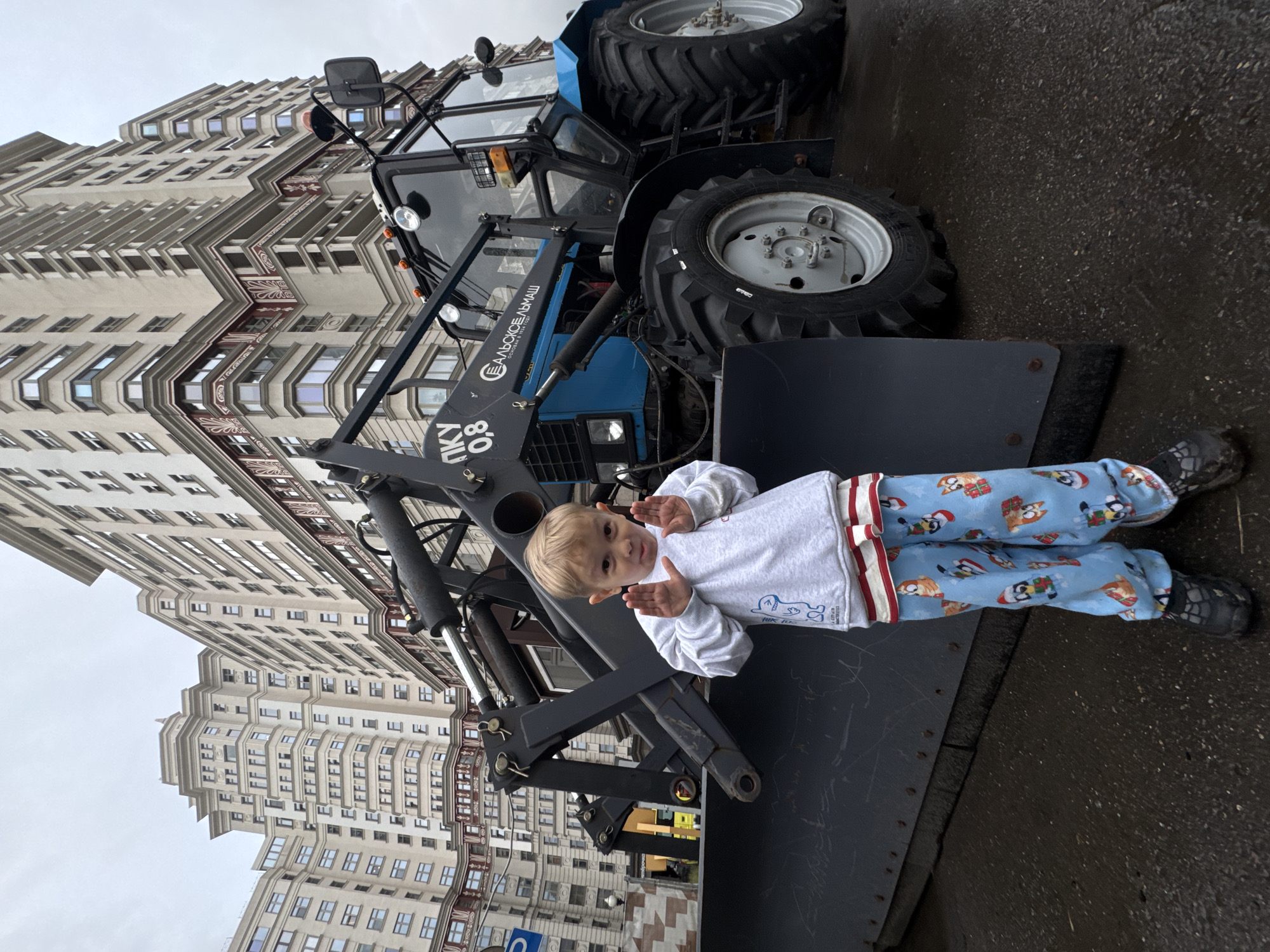 A young boy stands in front of a blue tractor with a snowplow attachment on a cloudy day. The boy is wearing a grey sweatshirt and blue pajamas with a pattern of cartoon dogs wearing Santa hats. The tractor is large and appears to be parked on a wet asphalt surface, likely after a rain. In the background, several large, ornate residential buildings, characteristic of Stalinist architecture, are visible. These buildings suggest the location is in Moscow, Russia, possibly on a street near residential areas. The overall scene appears to be a public space, given the presence of the tractor and the large buildings.