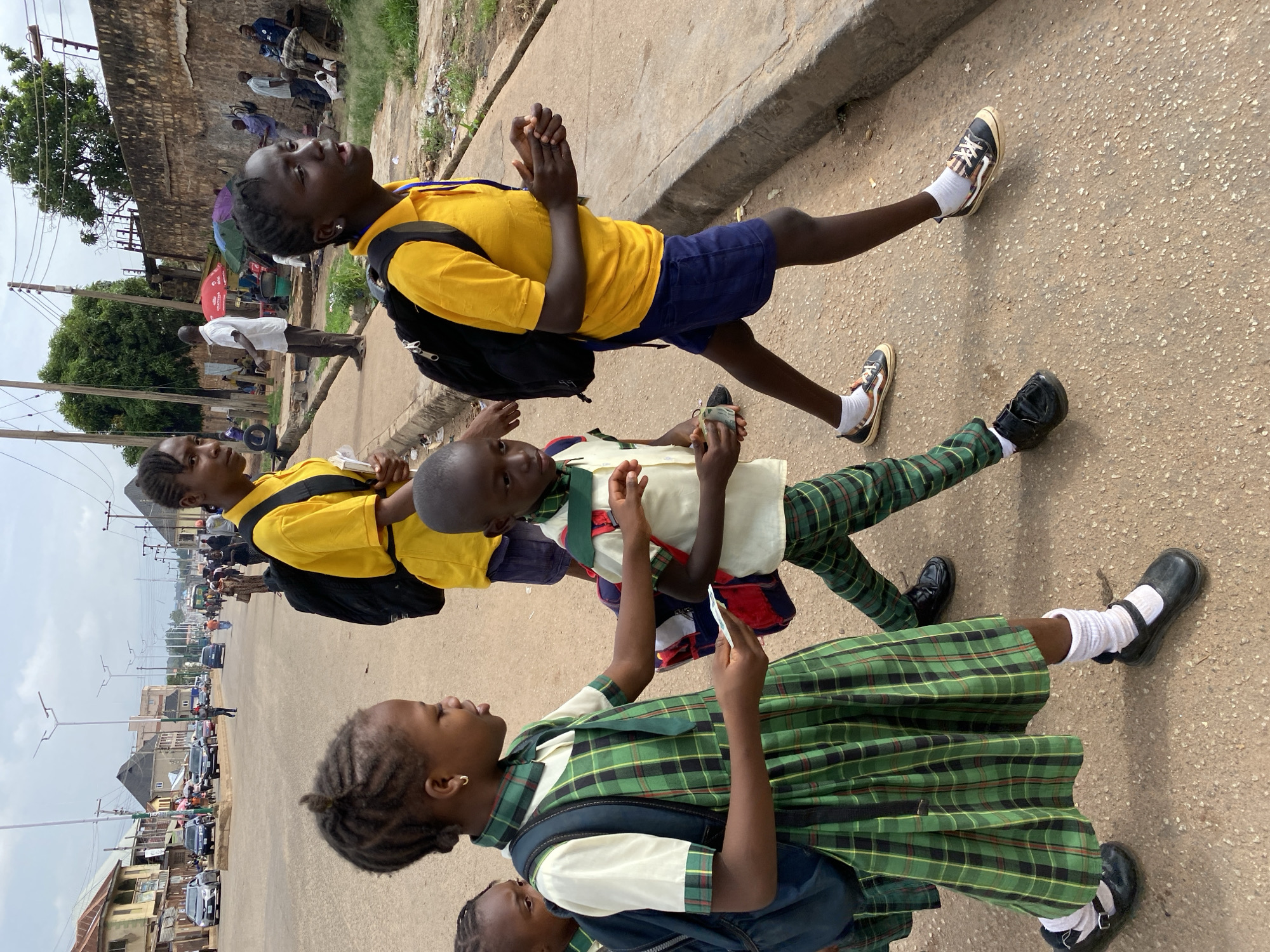 In Bukuru, Nigeria, a group of individuals, predominantly children in school uniforms, walk along a paved road. The foreground is dominated by four children. A girl on the left, wearing a green and black plaid dress with a white collar and cuffs, a green tie, and black Mary Jane shoes with white socks, carries a dark blue backpack. She is looking down at a small white object in her hand. To her right, a young boy in a light-colored shirt with a green collar and cuffs, green and black plaid pants, and black shoes with white socks, walks with a blue and red backpack slung over his shoulder. He holds a small stack of what appear to be currency notes in his hands. Behind them, a teenager with braided hair, wearing a yellow t-shirt and dark blue shorts, walks with a black backpack. To the far right, another teenager in a yellow t-shirt and dark blue shorts walks with a black backpack. The road appears to be a busy thoroughfare with other pedestrians and vehicles in the background, including cars and what might be a bus. Buildings line the left side of the road, and a concrete drainage channel runs along the right side, with a stone wall beyond it. The sky is overcast.