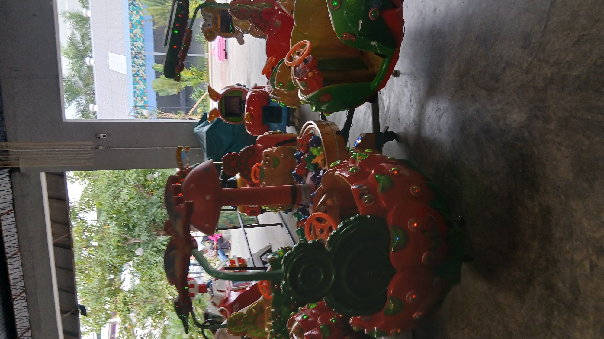 In an indoor, covered open-air structure with a grey concrete floor and a metal roof structure, a variety of colorful, child-sized amusement rides are arranged. The rides, designed to resemble fruits and cartoon characters, feature bright red, green, and yellow components, with some equipped with orange steering wheels. In the background, through open doorways, glimpses of trees, a grey building with a pitched roof, and what appears to be a water feature with blue tiling are visible, suggesting an outdoor area connected to the indoor space. A few individuals are seated in the background, observing the amusement rides. The overall scene depicts an amusement area likely located in the City of Medan, Indonesia, characterized by its playful and vibrant design.