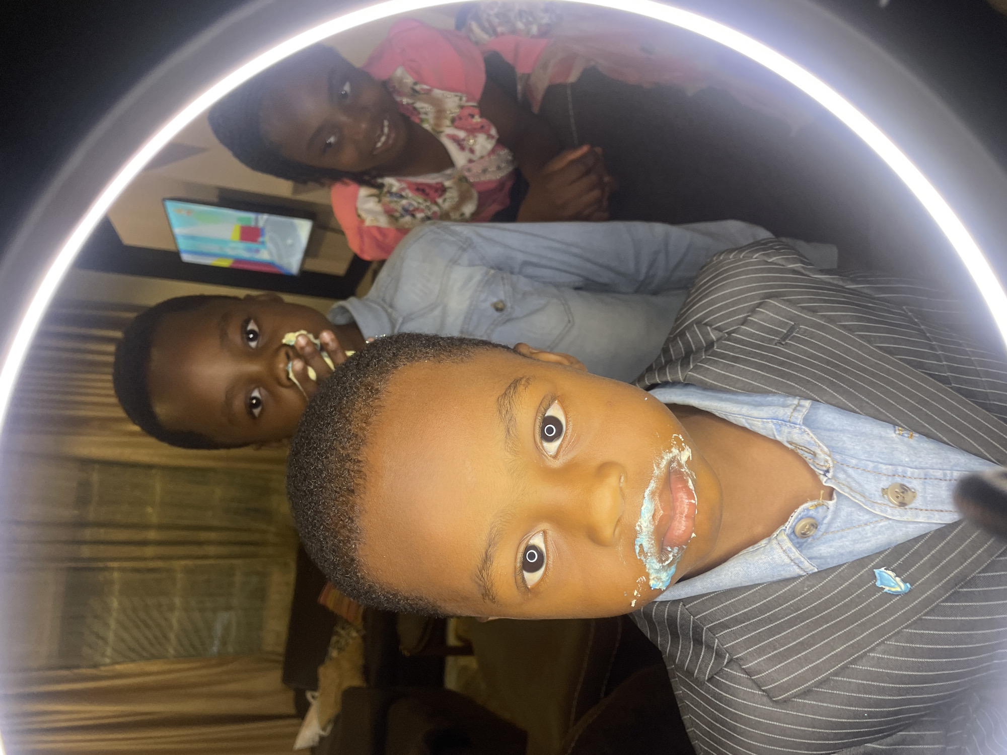 Three children are captured in a photo booth-style image with a ring light illuminating the scene.
The foreground features a young boy dressed in a pinstripe blazer over a denim shirt. He has frosting or cake smeared around his mouth.
Behind him, another boy with similar frosting on his face leans back, wearing a denim shirt.
In the background, a girl is seen in a floral patterned shirt.
A television screen is visible in the upper left, displaying multicolored abstract shapes.
The location is unknown, and there's no definitive information indicating it is Jos, Nigeria.