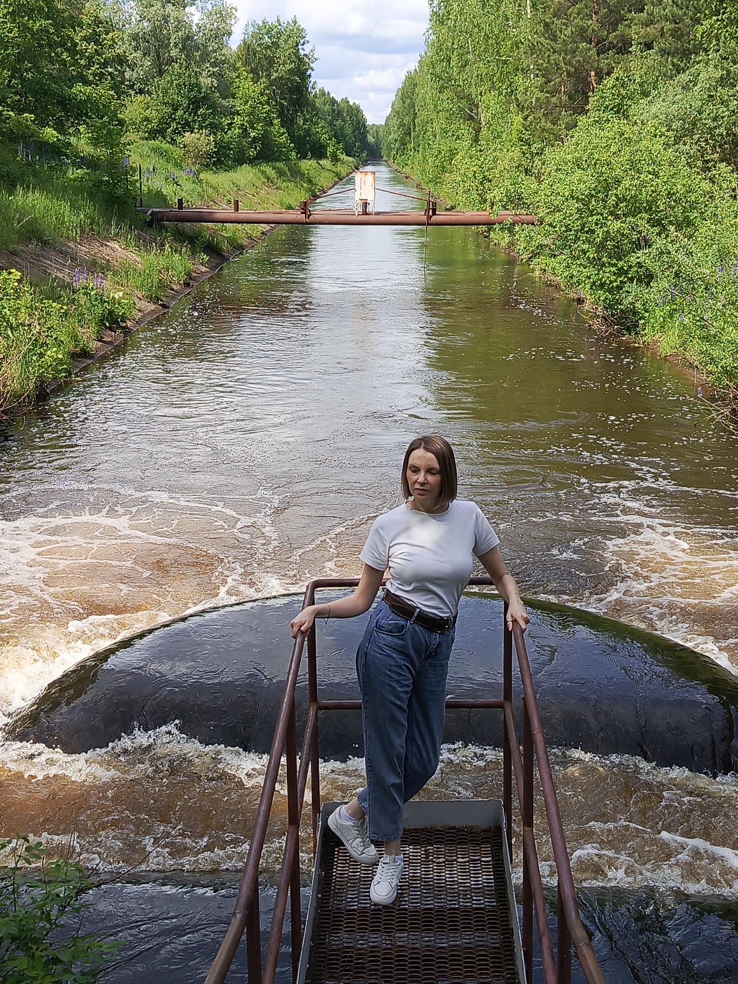 A woman stands on a metal platform with railings, looking slightly to her right, in an outdoor setting. She has short, light brown hair and is wearing a white short-sleeved t-shirt, blue jeans with a brown belt, and white sneakers. Her expression is neutral.
The platform she is standing on overlooks a section of water that appears to be flowing over a drop or weir, creating foamy, brownish-white rapids directly below her. Beyond this turbulent section, the water of a canal or river flows calmly. The water in the calmer sections has a murky, brownish-green hue.
The canal is lined on both sides by lush green vegetation, including tall trees and bushes, suggesting a natural or semi-natural environment. A rusty metal pipe crosses over the canal further down, supported by vertical structures on the banks. On the banks, patches of purple flowers, possibly lupines, are visible among the green grass.
The scene is illuminated by bright daylight, indicating it is daytime, likely mid-morning or early afternoon, under a clear or partly cloudy sky. The overall impression is one of a serene natural landscape with an industrial or man-made element (the platform, pipe, and water management structure) integrated within it. No visible text is present in the image.