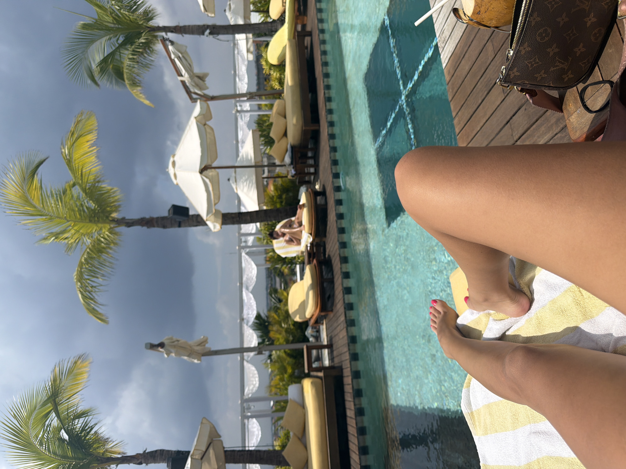 The image presents a first-person perspective of an outdoor poolside lounge area in Tibubeneng, Indonesia. In the immediate foreground, the bare, tanned legs and feet of an individual, displaying pink toenail polish, are visible, resting on a yellow and white striped towel. To the right of the legs, on a wooden deck, are a brown Louis Vuitton Monogram Pochette Accesoires bag, a black elastic hair tie, and a natural coconut drink with a white straw.
The midground features a clear blue swimming pool with discernible square mosaic tiles at its bottom, bordered by a wooden deck and a dark checkered tile edge. Multiple yellow upholstered sun loungers and daybeds are arranged around the pool. One individual is observed reclining on a yellow chaise lounge on the far side of the pool, partially obscured. Several white patio umbrellas are open, and mature palm trees are interspersed throughout the scene.
The background reveals an overcast sky with dark grey clouds. A white railing adorned with draped white fabric or netting is visible beyond the pool, with indistinct structures present on the distant horizon.