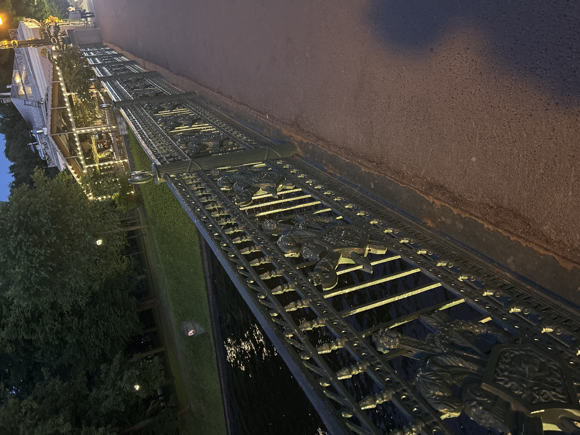 In the city of Unknown, Unknown, a bridge railing with intricate metalwork and decorative figures overlooks a dark body of water. Beyond the water, a grassy area is bordered by trees. Further in the background, a building with a glass exterior and illuminated by numerous lights stands prominently. The building appears to be a restaurant or cafe, with people visible inside. A paved walkway runs alongside the bridge railing, and a street lamp emits a warm glow. The scene is captured at dusk or night, with the sky a deep blue and the lights casting long shadows. One person is walking on the walkway in the distance.