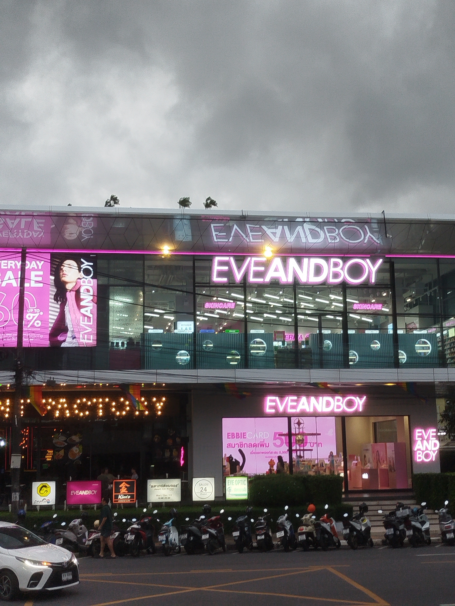 This media file captures a street-level view of a commercial building, likely a shopping center or retail store, on a cloudy day. The prominent signage indicates the presence of a store named "EVEANDBOY," which appears to be a cosmetics or beauty retailer. This name is displayed in large, illuminated pink lettering on multiple levels of the building.
Below the main EVEANDBOY signage, a large digital display features a woman with dark hair looking to her left, with text that appears to be in Thai and English, promoting a sale and possibly a membership program (indicated by "EBBIECARD" and "สมาชิกลดเพิ่ม" which translates to "member discount increase"). Other signs in Thai and English are also visible, suggesting a diverse clientele or international presence.
In front of the building, a row of parked motorcycles suggests a busy urban environment. A white car is parked on the left side of the frame, and a person is walking between the car and the motorcycles. The overall scene suggests a typical day in a commercial district, with overcast skies indicating either early morning, late afternoon, or impending rain. The lighting from the signs provides a bright contrast to the subdued natural light. The presence of rainbow-colored banners above the "FACTORY" sign hints at a possible Pride event or a business associated with the LGBTQ+ community. The building itself has a modern facade with large glass windows reflecting the interior and the sky.
