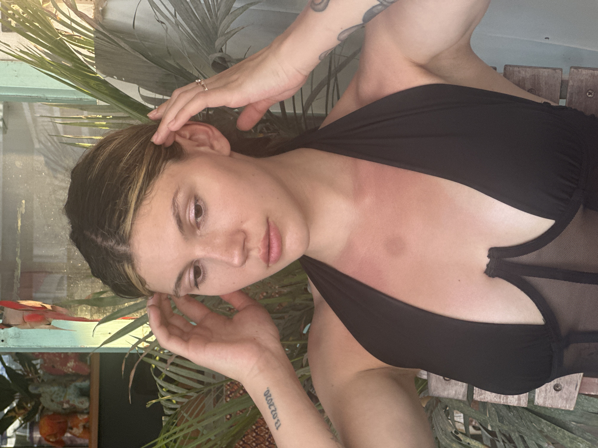 A woman is facing the camera with her head tilted to the left. She is wearing a black top with mesh detailing. Her hair is dark with light highlights and is styled back. She has tattoos on both arms, one clearly showing the date "13.02.2020". Her hands are near her ears. The background contains foliage, including palm-like leaves. A structure with a blue-painted frame is visible behind her. The scene takes place outdoors in a location that is presumably Baan Bang Rak, Thailand.