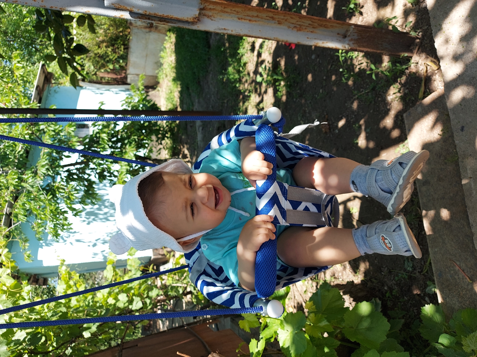 A baby sits in a blue and white striped swing under a metal frame structure. The baby is wearing a white hat, a blue shirt, and gray sandals with matching socks. The child is smiling and appears to be enjoying the swing. Green foliage surrounds the swing, and a light blue building is partially visible in the background. The location is Tavaksay, Uzbekistan, based on the information provided.