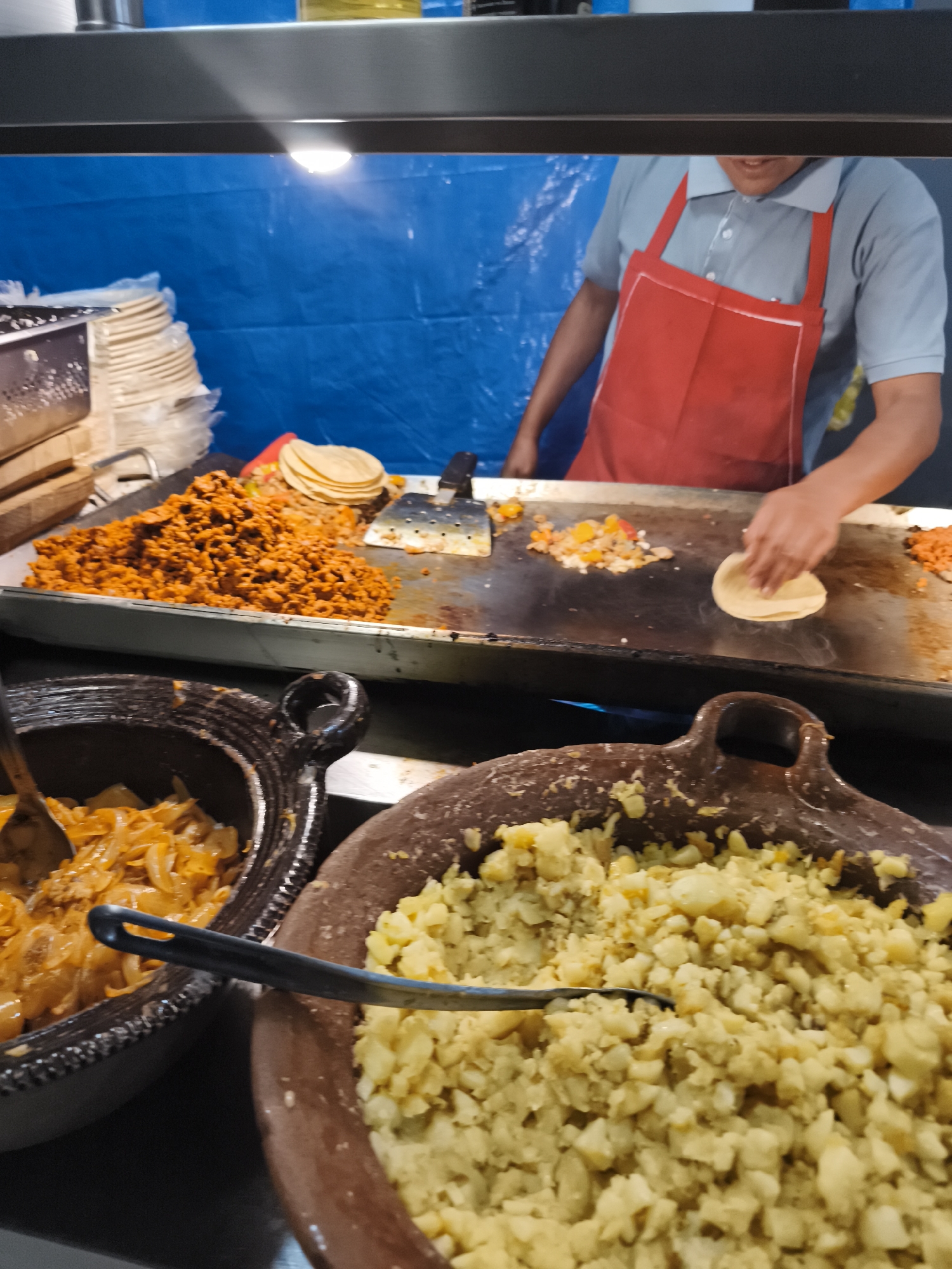 The photograph shows a food vendor in Mexico City preparing tacos. The vendor, wearing a red apron and light blue shirt, is heating tortillas on a griddle that also contains chopped vegetables and seasoned meat. To the left of the griddle is a stack of tortillas. In the foreground are two large clay pots containing cooked potatoes and sautéed onions. Utensils are present in each pot.